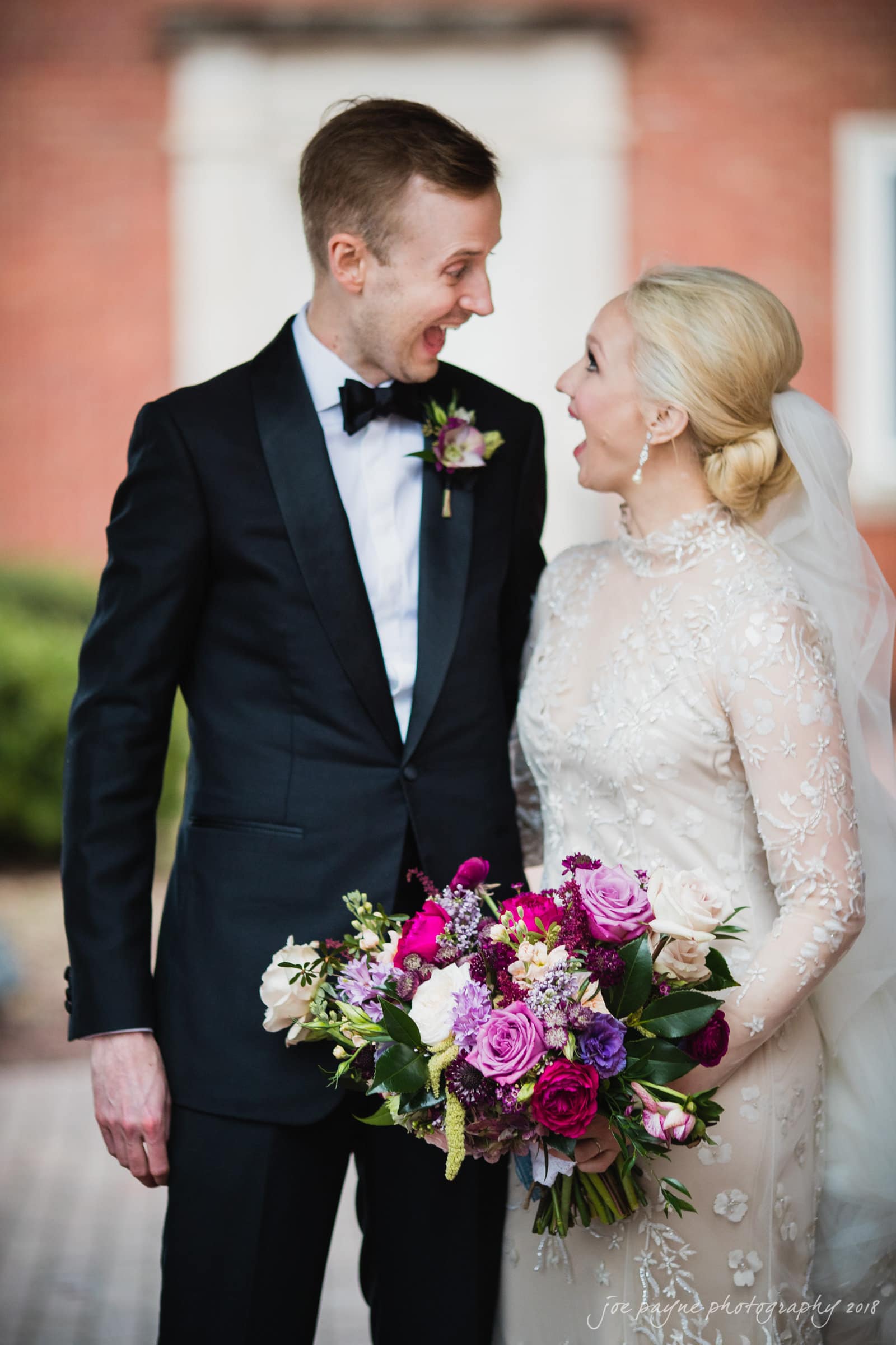 st. mary's & umstead wedding photographer - sarah & jake A groom in a black tuxedo and a bride in a white lace dress share a joyful moment, each with surprised expressions. The bride holds a vibrant bouquet of roses and greenery. They stand outdoors with a blurred brick building in the background.