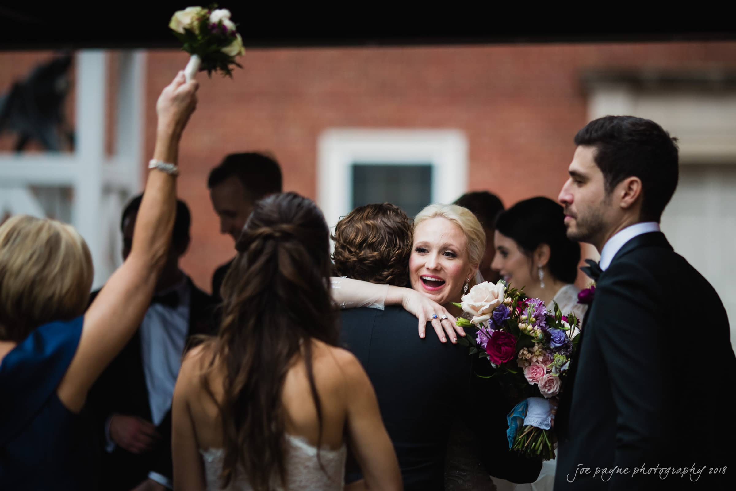 st. mary's & umstead wedding photographer - sarah & jake A joyful wedding scene with a smiling bride embracing a guest. She holds a bouquet of vibrant flowers. Other well-dressed guests are around, one woman raising a small bouquet. They stand against a blurred brick background.