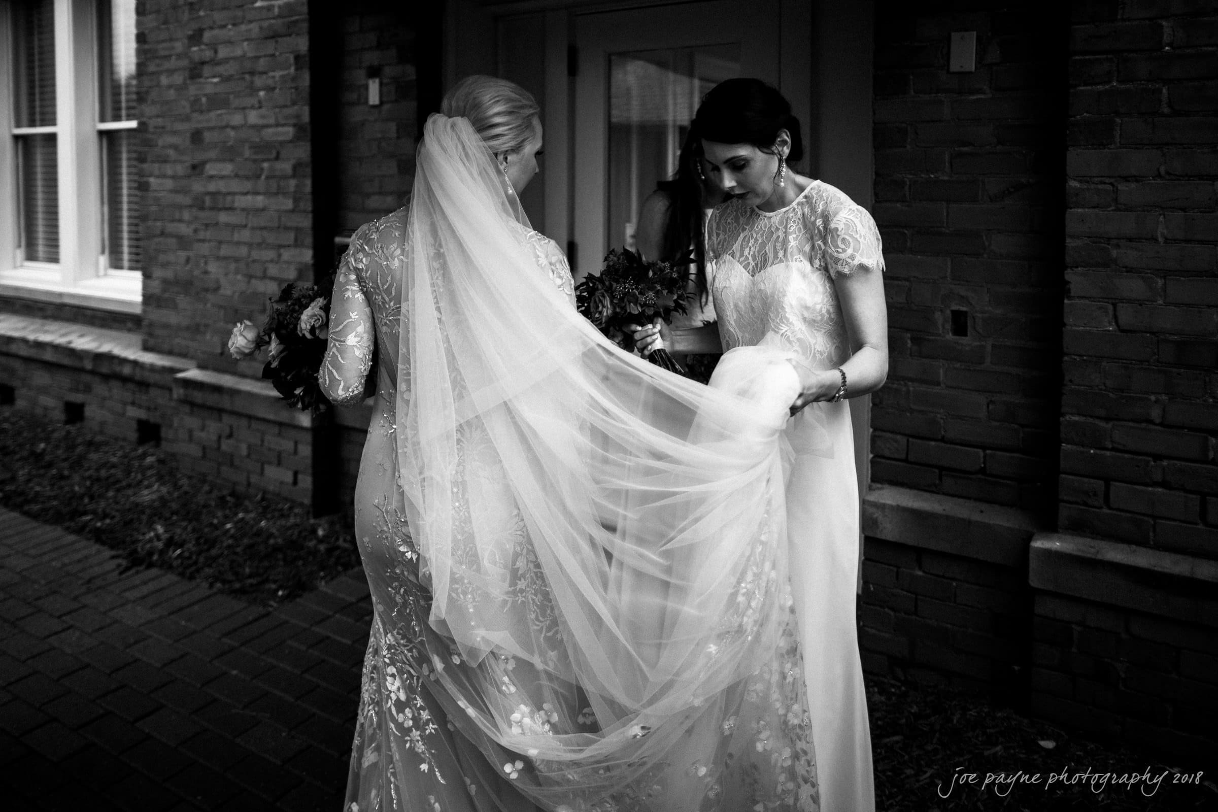 st. mary's & umstead wedding photographer - sarah & jake Black and white photo of two women in lace bridal gowns standing outside a building. One holds a bouquet, while the other adjusts the long veil and train of the dress. They appear engaged in conversation.