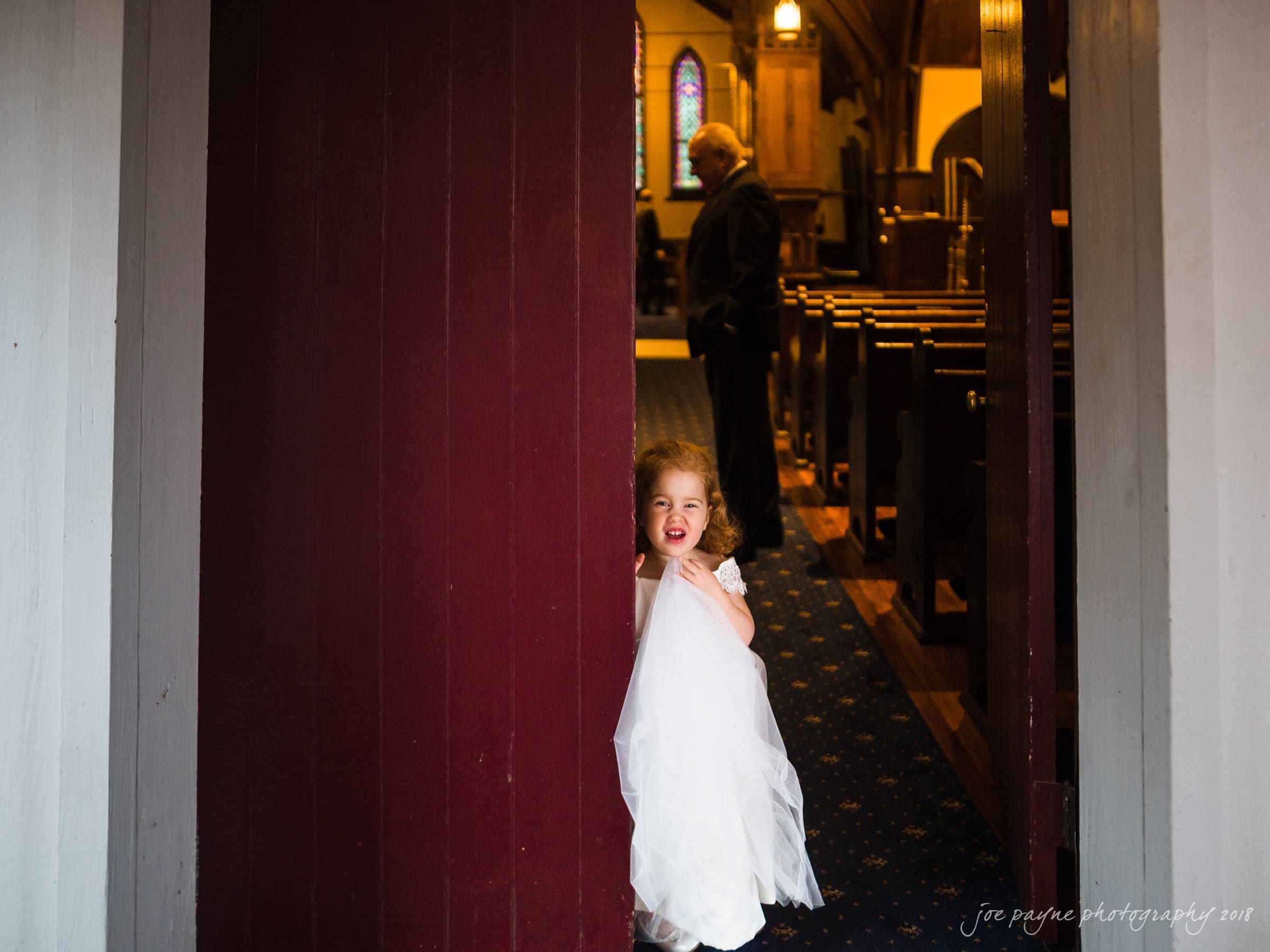 st. mary's & umstead wedding photographer - sarah & jake A young girl in a white dress stands smiling in a church doorway. She is partially hiding behind a red wall. The interior of the church is softly lit, with wooden pews and a stained glass window visible in the background.