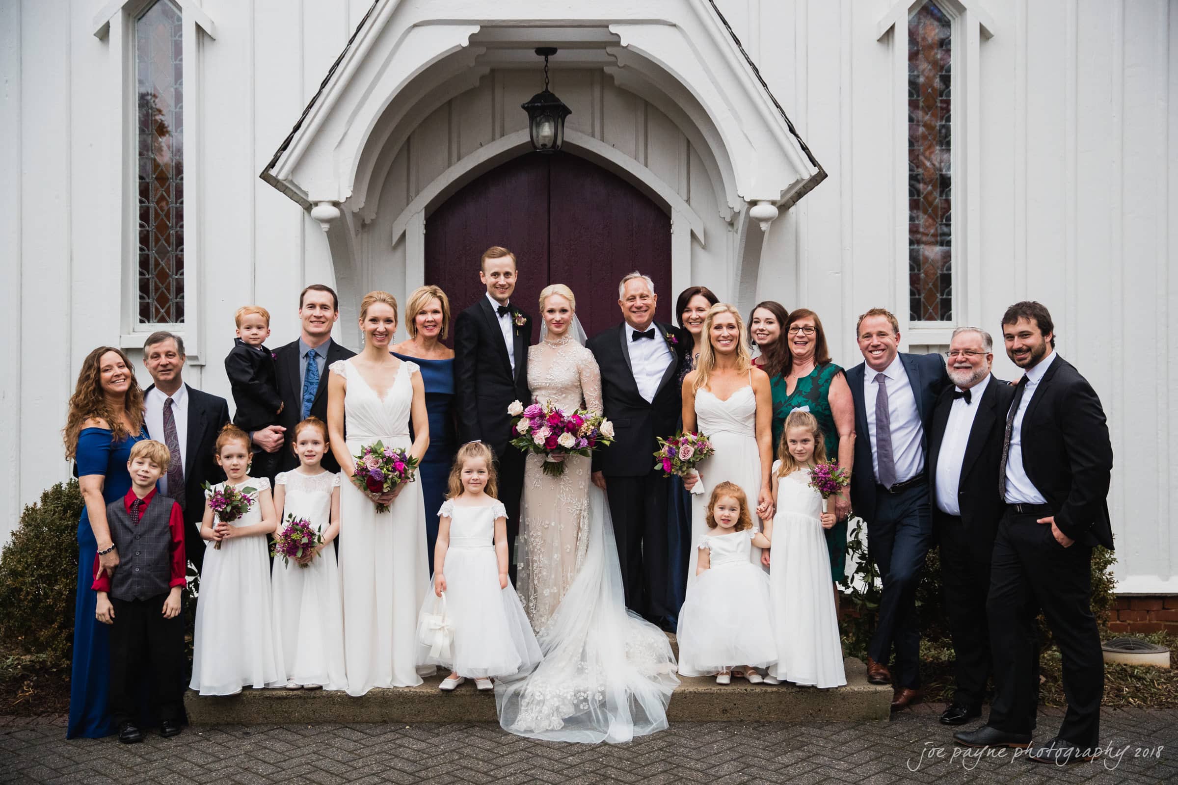 st. mary's & umstead wedding photographer - sarah & jake A wedding party poses in front of a white church. The group includes the bride and groom, flower girls in white dresses, bridesmaids in lavender, groomsmen in suits, and family members. Everyone is smiling and holding bouquets.