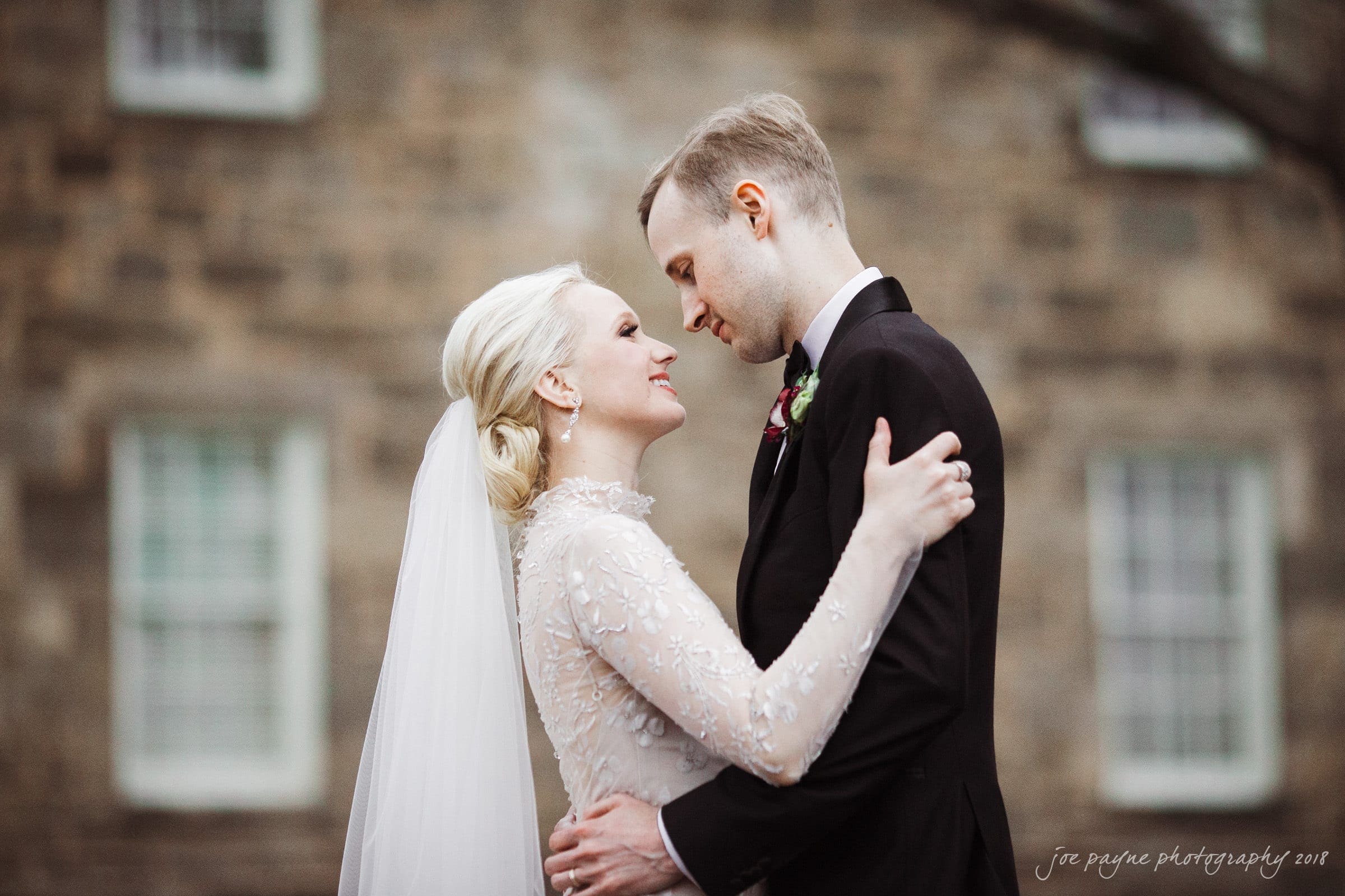 st. mary's & umstead wedding photographer - sarah & jake A bride and groom embrace and gaze at each other lovingly. The bride wears a white dress with lace details and a veil, while the groom is in a black suit. They are outdoors in front of a blurred brick building.