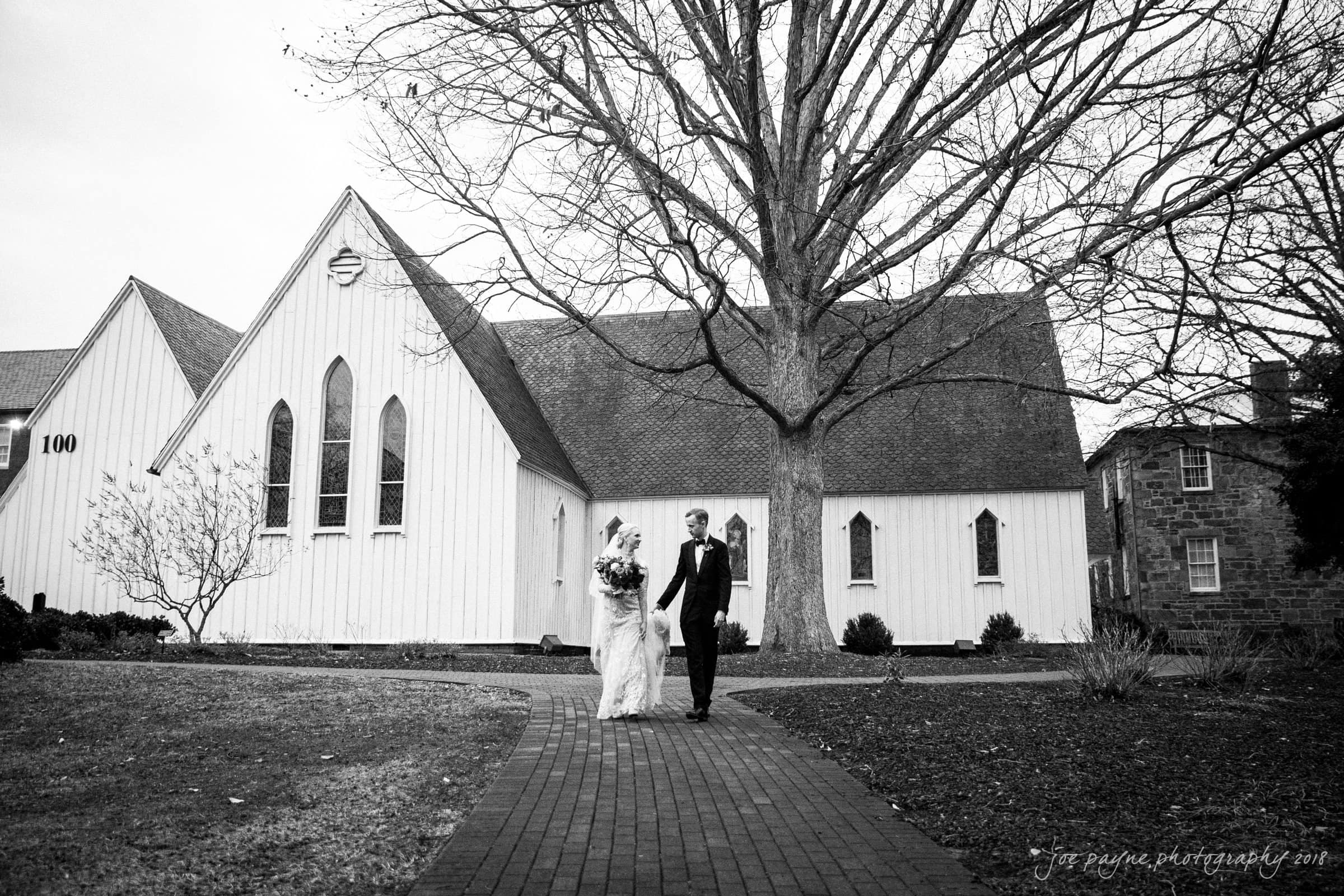 st. mary's & umstead wedding photographer - sarah & jake A bride and groom walk hand in hand on a brick path in front of a white chapel. The bride holds a bouquet, and bare trees surround the building. The scene is in black and white, capturing a timeless, serene moment.