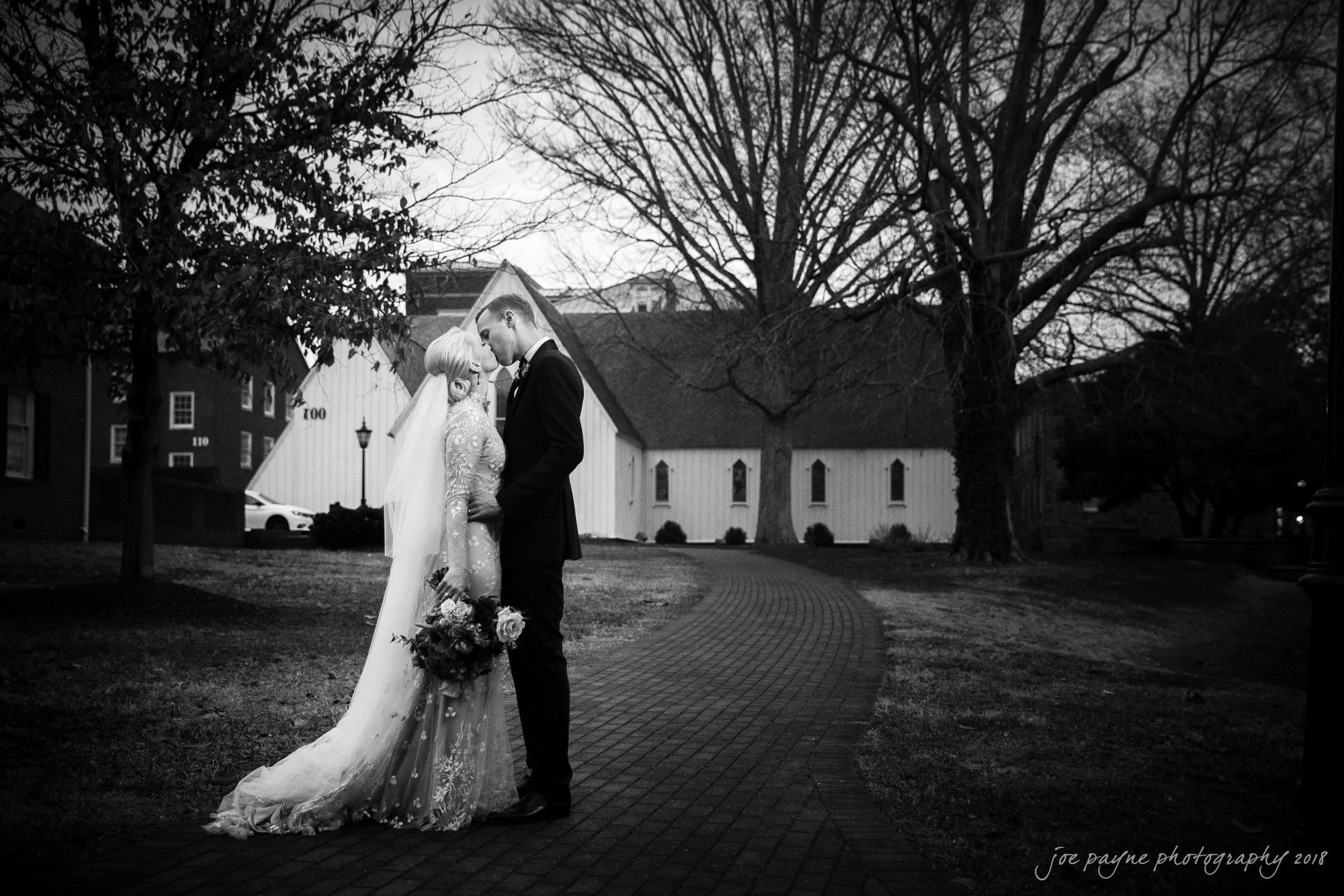 st. mary's & umstead wedding photographer - sarah & jake A black and white image of a bride and groom standing together on a brick path, sharing an intimate moment. The bride holds a bouquet, and they are surrounded by trees and a historic building in the background.