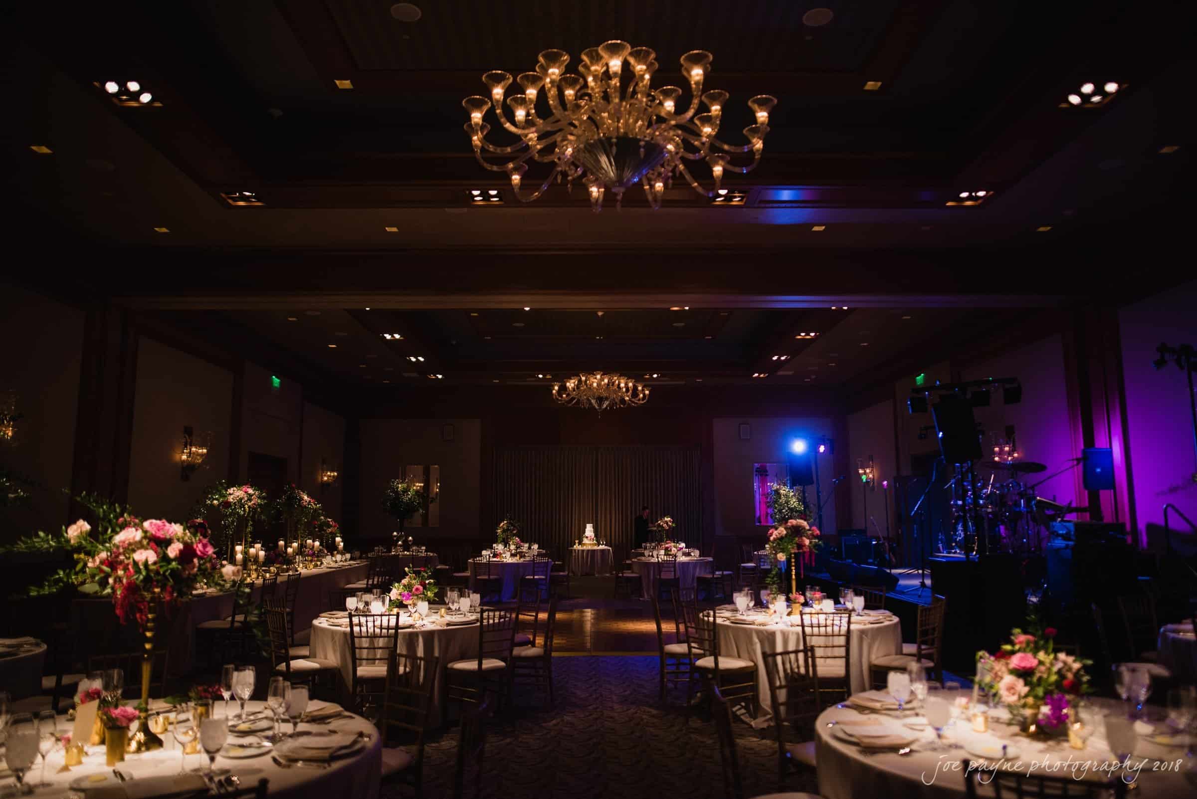 st. mary's & umstead wedding photographer - sarah & jake A beautifully decorated wedding reception hall with dim lighting. Elegant chandeliers hang from the ceiling, and tables are set with floral centerpieces and white table settings. A small cake is spotlighted in the center of the room.