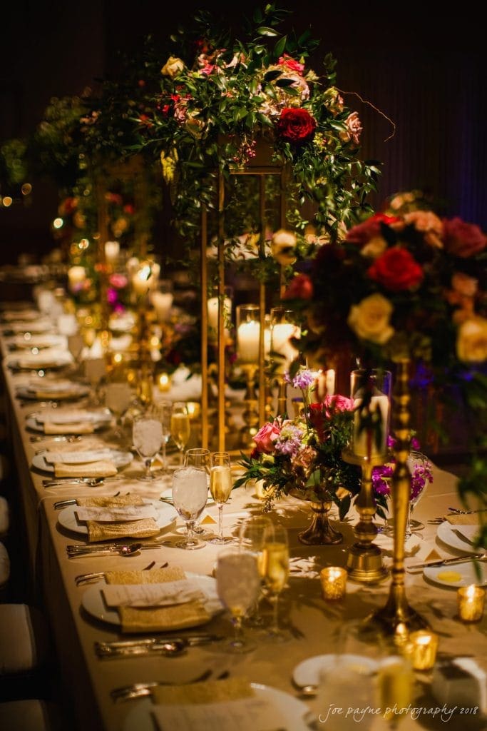 A beautifully set wedding reception table adorned with elegant floral centerpieces, gold candle holders, and sparkling glassware. Warm lighting casts a romantic glow over the table, creating an inviting and celebratory atmosphere.