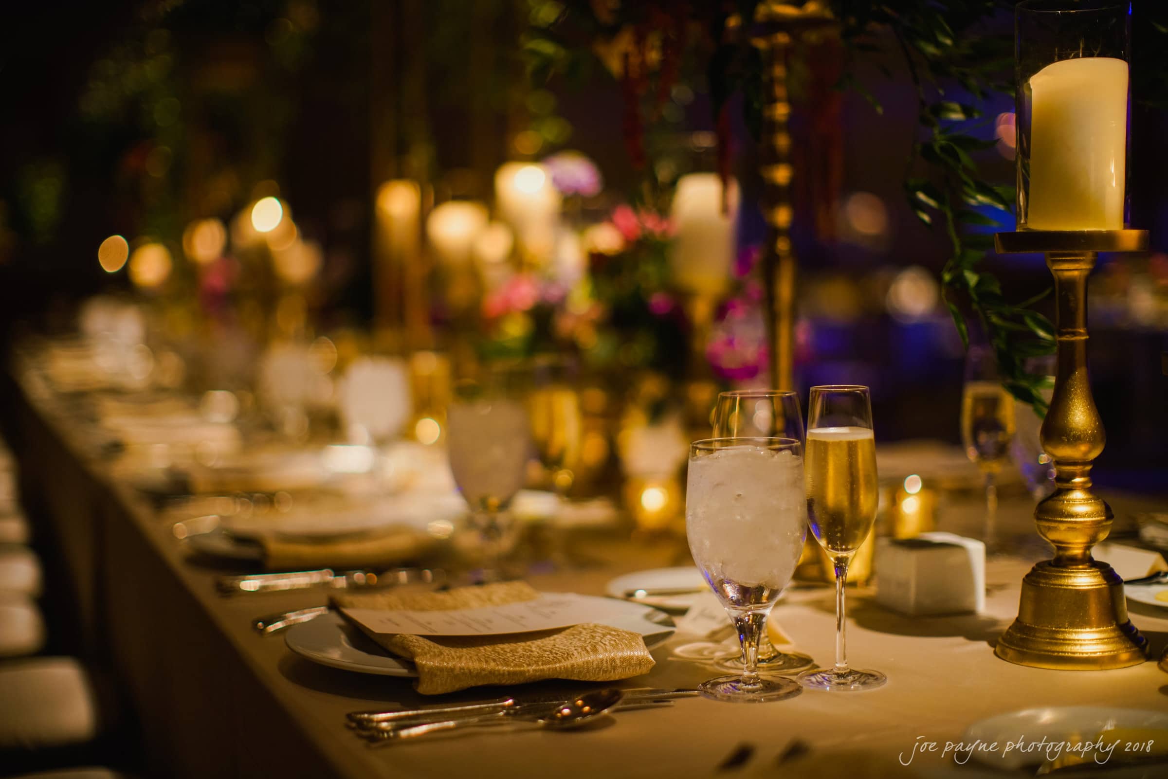 st. mary's & umstead wedding photographer - sarah & jake A beautifully set dining table with elegant gold candlesticks, lit candles, and crystal glassware. The dim lighting creates a warm, intimate atmosphere. Plates with napkins and menus are arranged neatly along the table.
