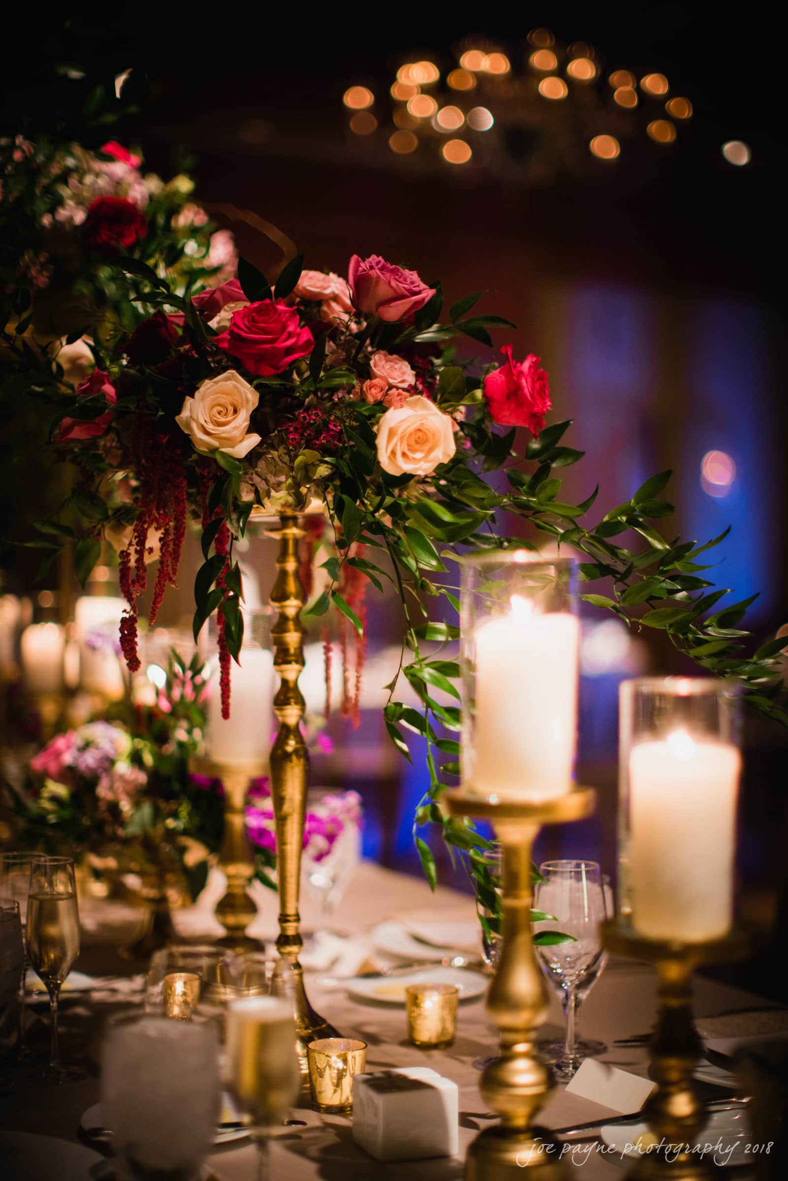 st. mary's & umstead wedding photographer - sarah & jake A lavish table setting featuring tall gold candle holders and a floral centerpiece with red, pink, and cream roses. Soft candlelight illuminates the elegant scene, creating a warm and romantic atmosphere.