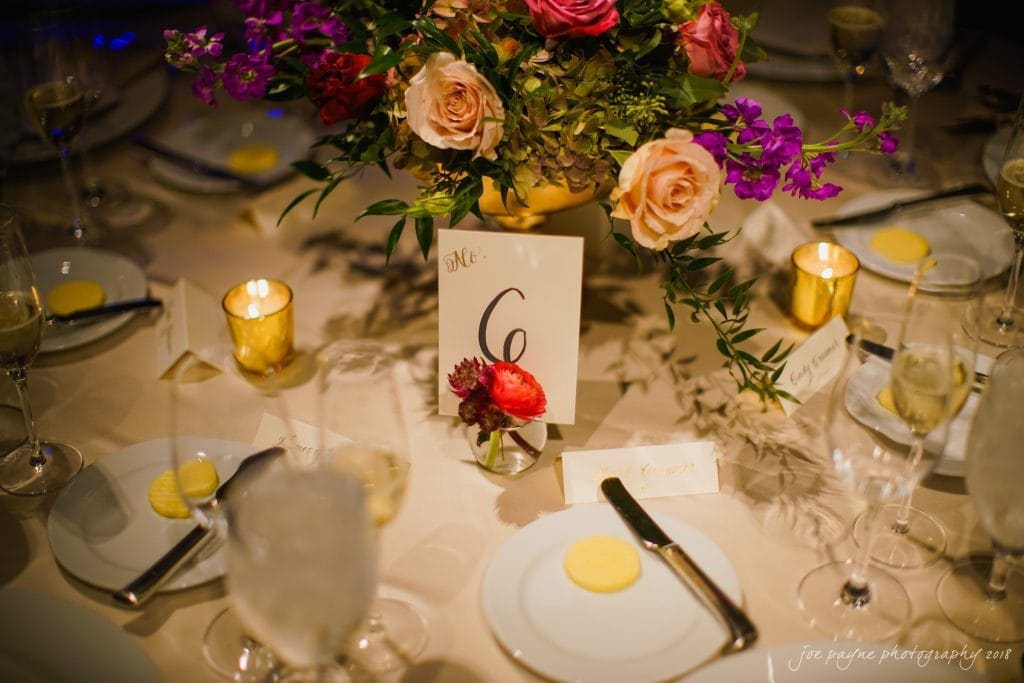 A beautifully set dinner table with a centerpiece of pink and purple flowers. The table is marked with a card displaying the number 6. Plates with butter and fine glassware surround the centerpiece, illuminated by soft candlelight.