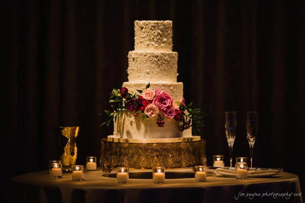 A four-tier white wedding cake adorned with pink and red flowers, placed on a gold stand. It is surrounded by lit candles and two champagne flutes on a round table, set against a dark background.