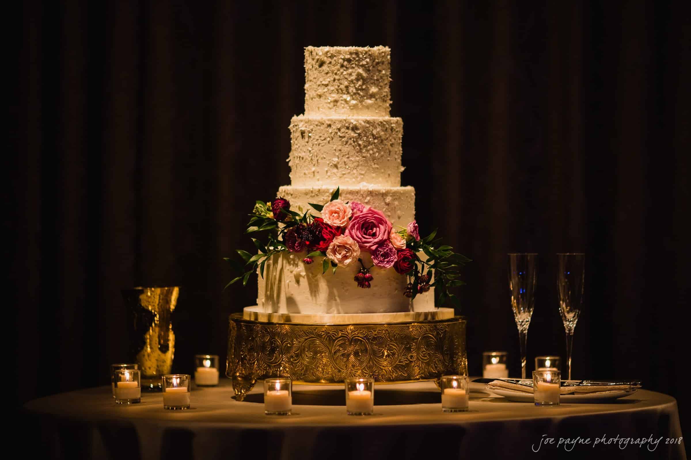 st. mary's & umstead wedding photographer - sarah & jake A four-tier white wedding cake adorned with pink and red flowers, placed on a gold stand. It is surrounded by lit candles and two champagne flutes on a round table, set against a dark background.