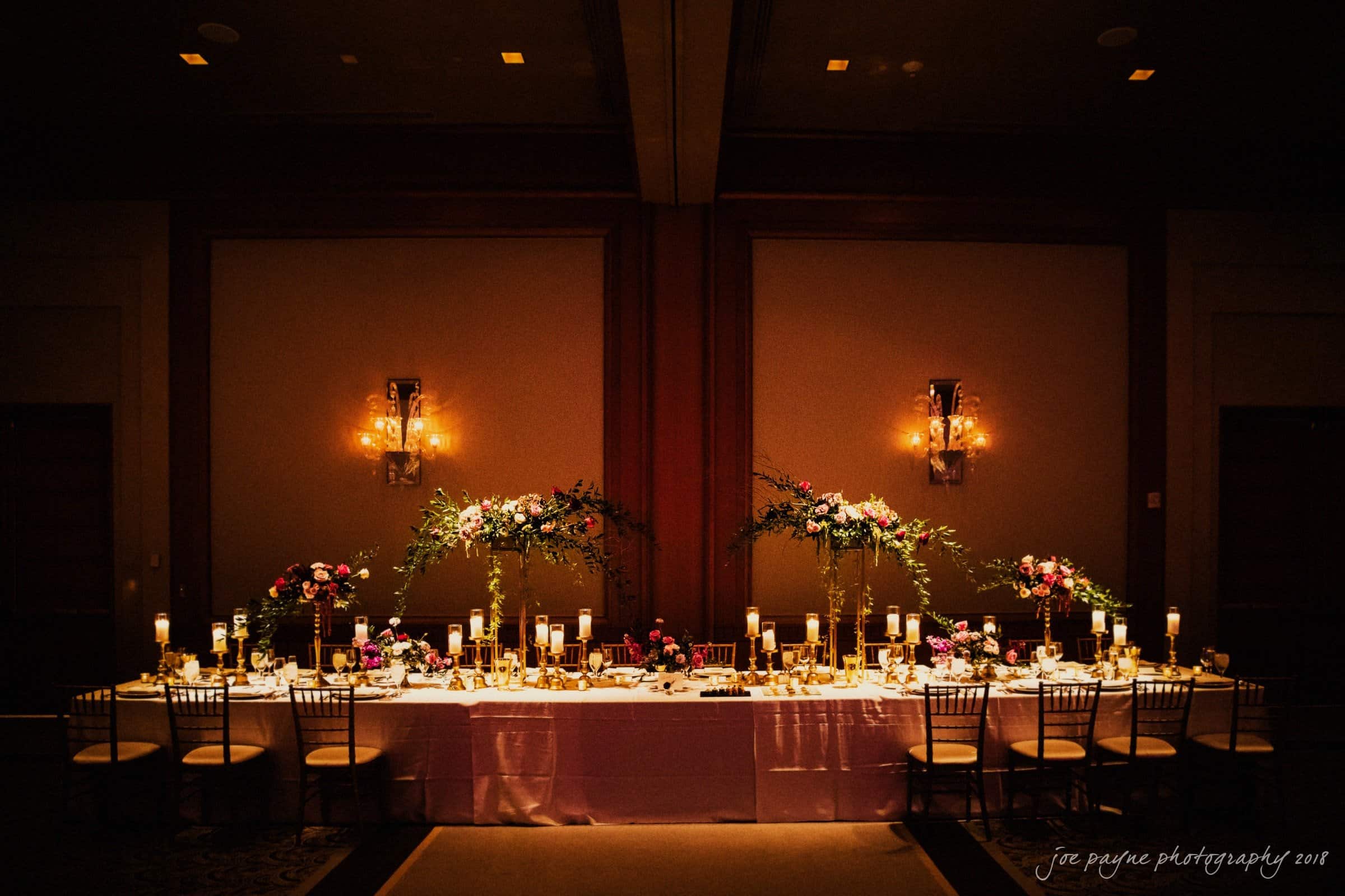 st. mary's & umstead wedding photographer - sarah & jake A beautifully decorated dining table set for a formal event. It features elegant floral centerpieces, lit candles, and neatly arranged tableware. The ambient lighting creates a warm and inviting atmosphere.