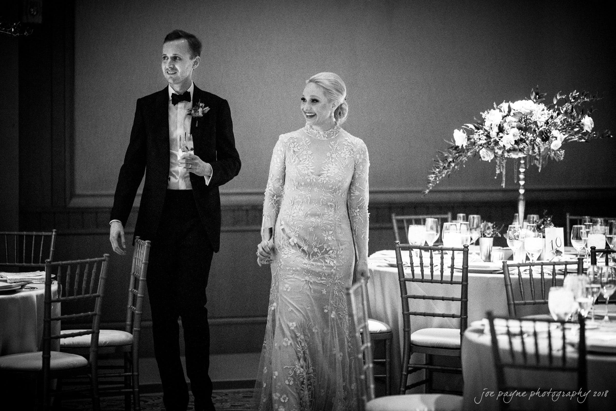 st. mary's & umstead wedding photographer - sarah & jake A couple, dressed elegantly, smiles and enters a beautifully decorated room with round tables and floral centerpieces. The woman wears a long gown, and the man is in a tuxedo. The atmosphere is formal and festive.