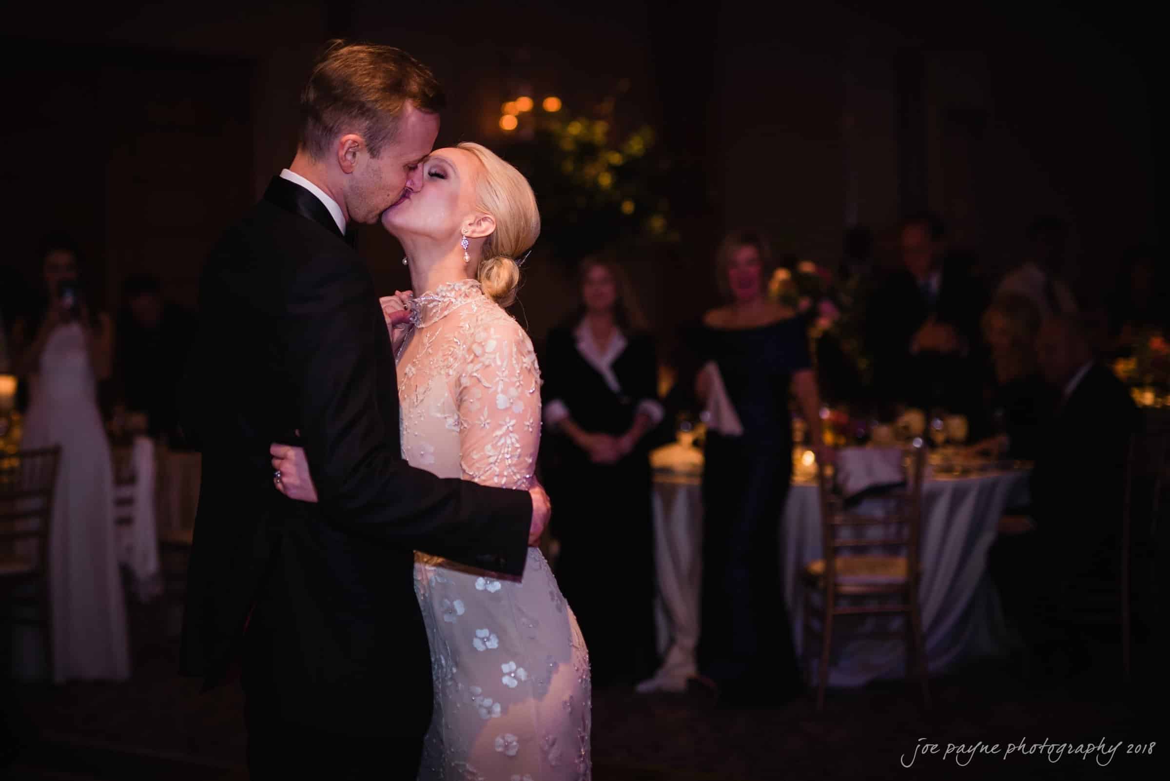 st. mary's & umstead wedding photographer - sarah & jake A bride and groom share a kiss on the dance floor during their wedding reception. The bride is wearing a lace dress with floral details, and the groom is in a black suit. Guests are seated at tables in the softly lit background.