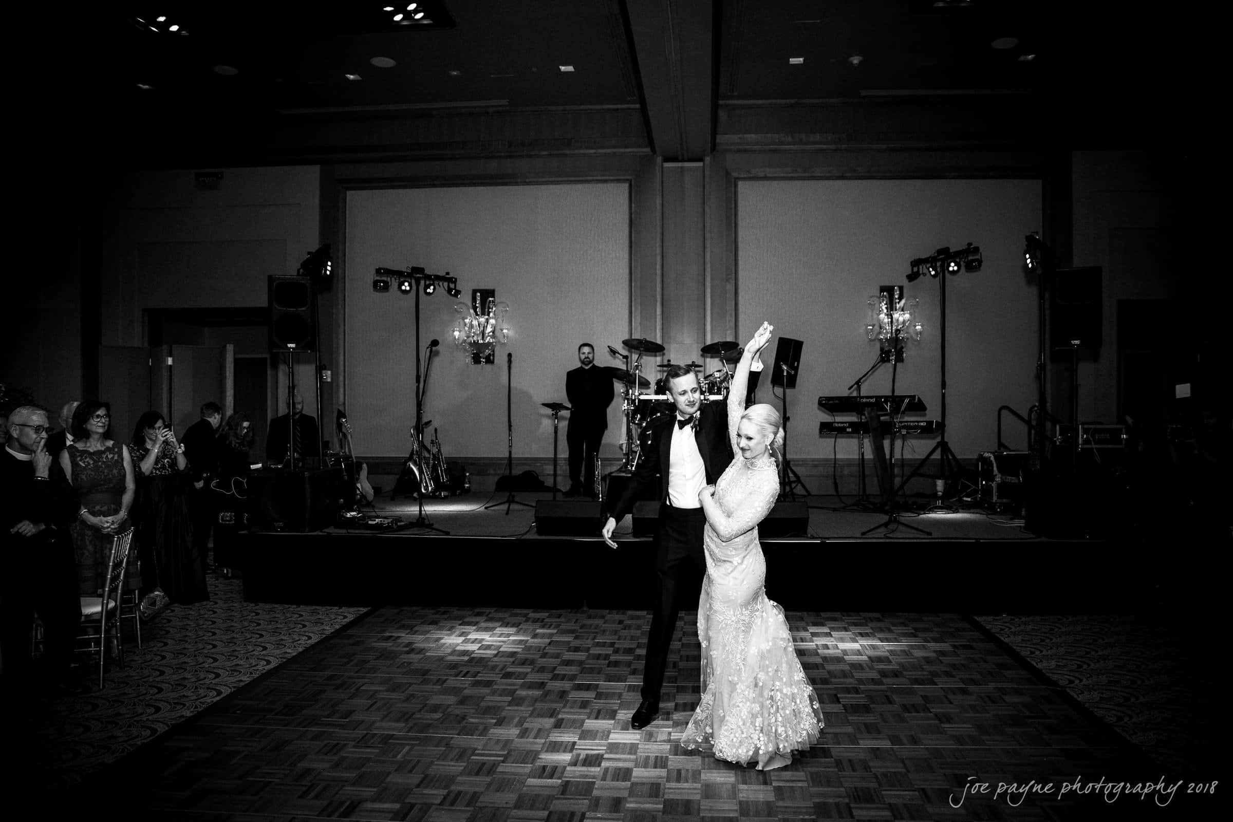 st. mary's & umstead wedding photographer - sarah & jake A couple elegantly dances on a wooden floor in a dimly lit ballroom, surrounded by an audience. Musical instruments and equipment are visible in the background, indicating a live band performance.