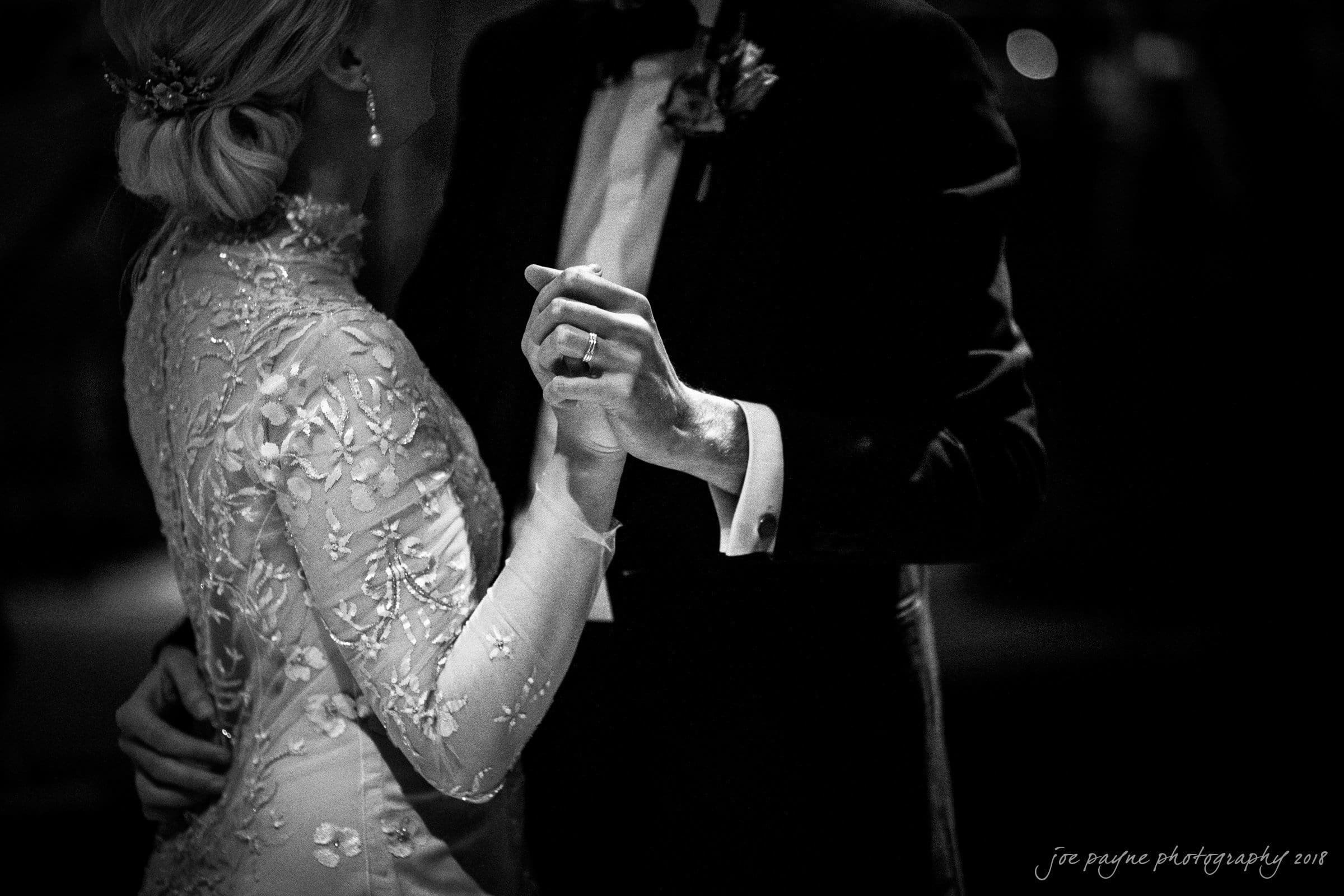 st. mary's & umstead wedding photographer - sarah & jake A black and white image of a couple dancing. The woman wears an embroidered dress and elegant earrings, while the man is in a suit. Their hands are clasped, and the scene is softly lit, creating a romantic atmosphere.