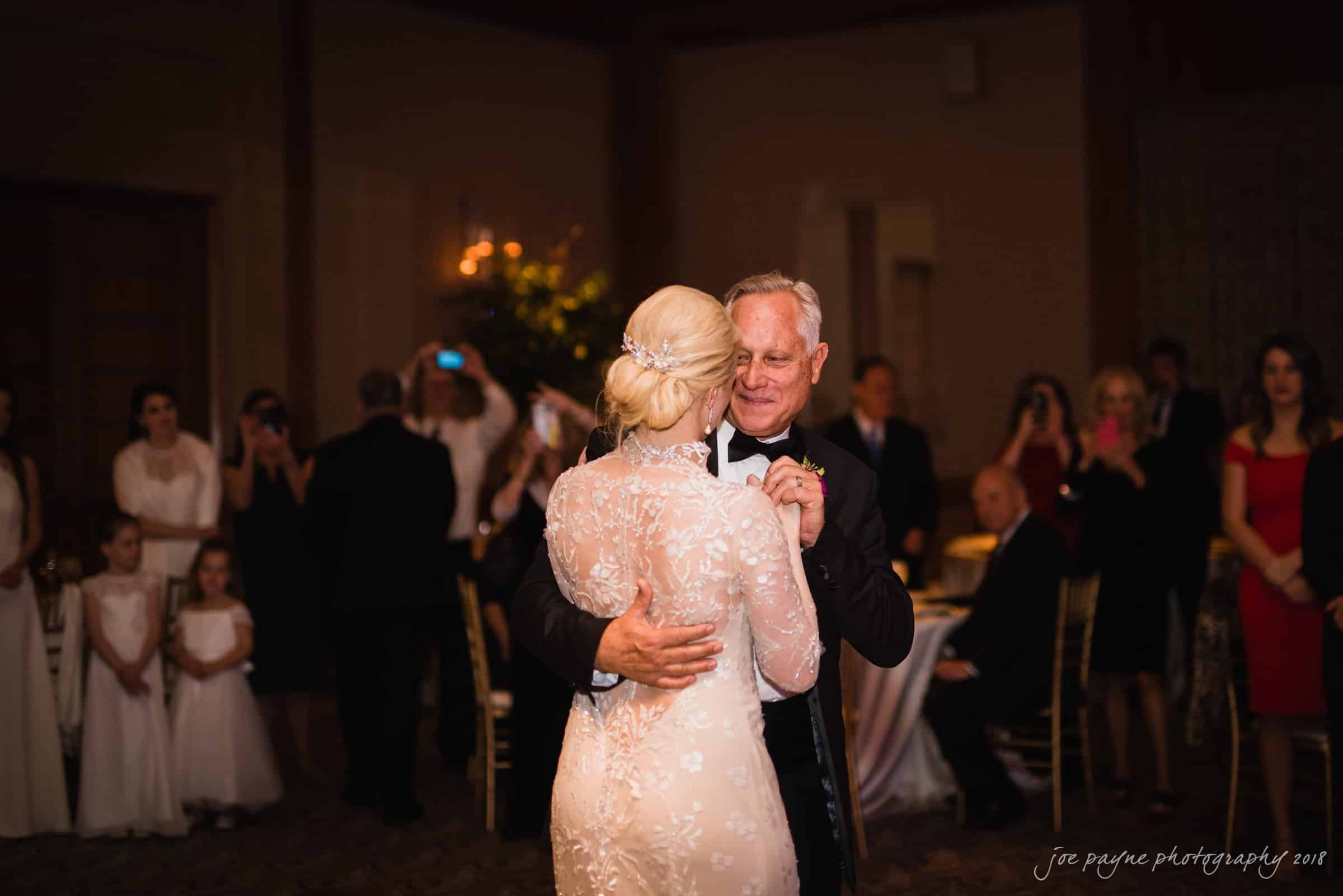 st. mary's & umstead wedding photographer - sarah & jake A bride and an older man share a dance at a wedding reception. They are surrounded by guests, some of whom are taking photos. The bride wears a white lace gown, and the setting is warmly lit with elegant decor.