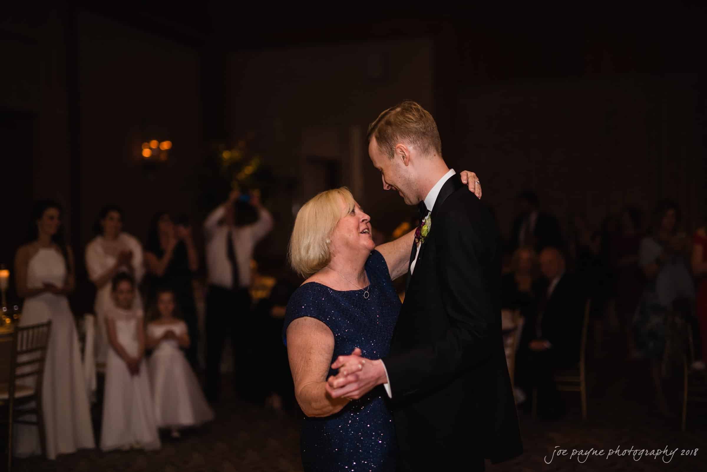 st. mary's & umstead wedding photographer - sarah & jake A man and woman are dancing in a warmly lit room, surrounded by seated and standing guests. The woman is smiling, and the man is wearing a suit. In the background, people are watching and taking photos.