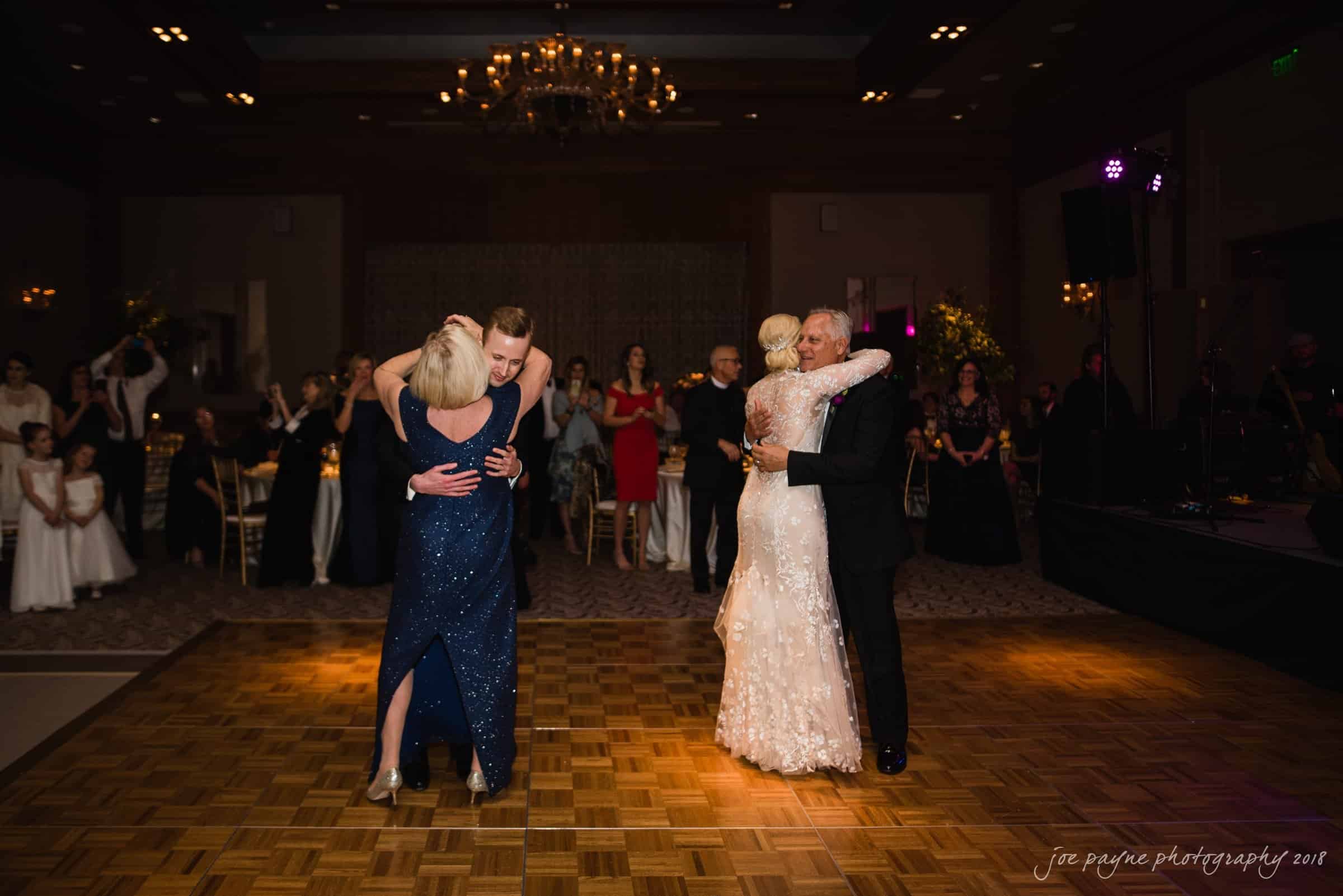 st. mary's & umstead wedding photographer - sarah & jake People dancing in pairs at a formal event on a wooden dance floor. The room is elegantly decorated with chandeliers and guests are seated in the background. The atmosphere is festive and joyful.