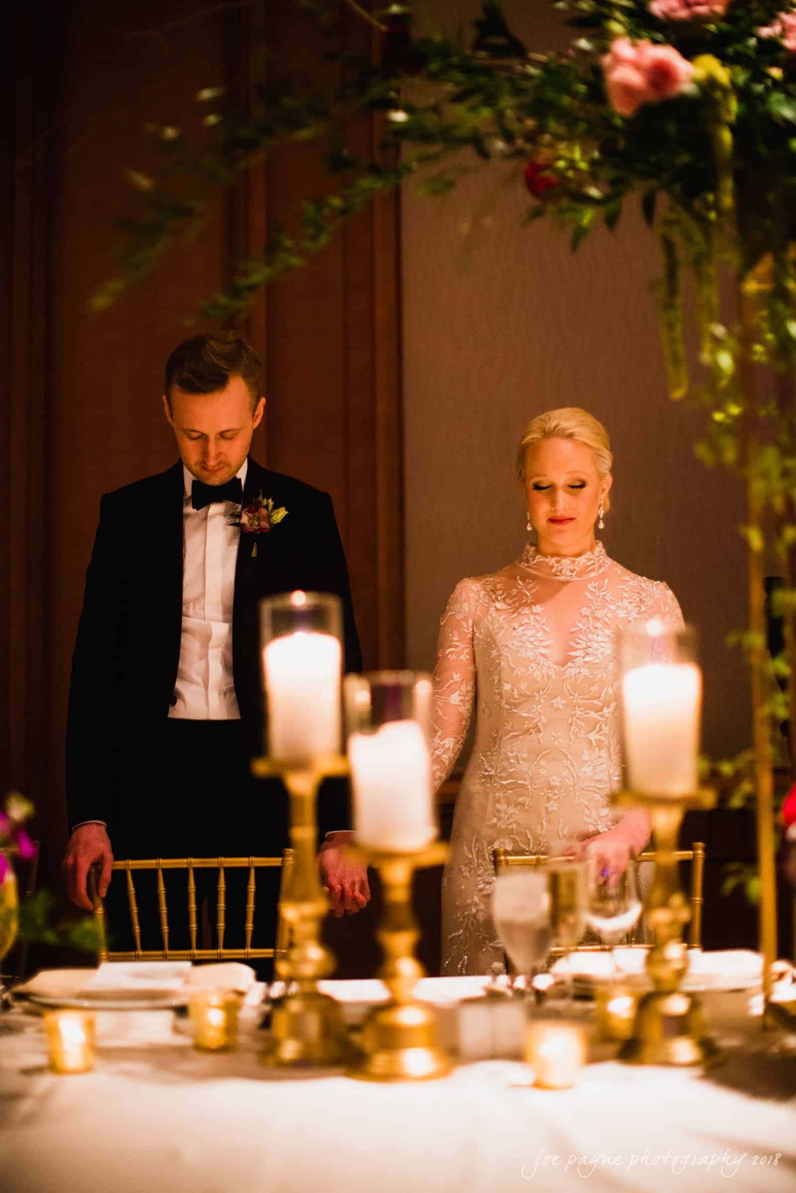 st. mary's & umstead wedding photographer - sarah & jake A couple stands holding hands at a decorated table during a formal event. The woman wears a detailed white dress, and the man is in a dark suit with a bow tie. Candlelight and floral arrangements create a warm, elegant atmosphere.