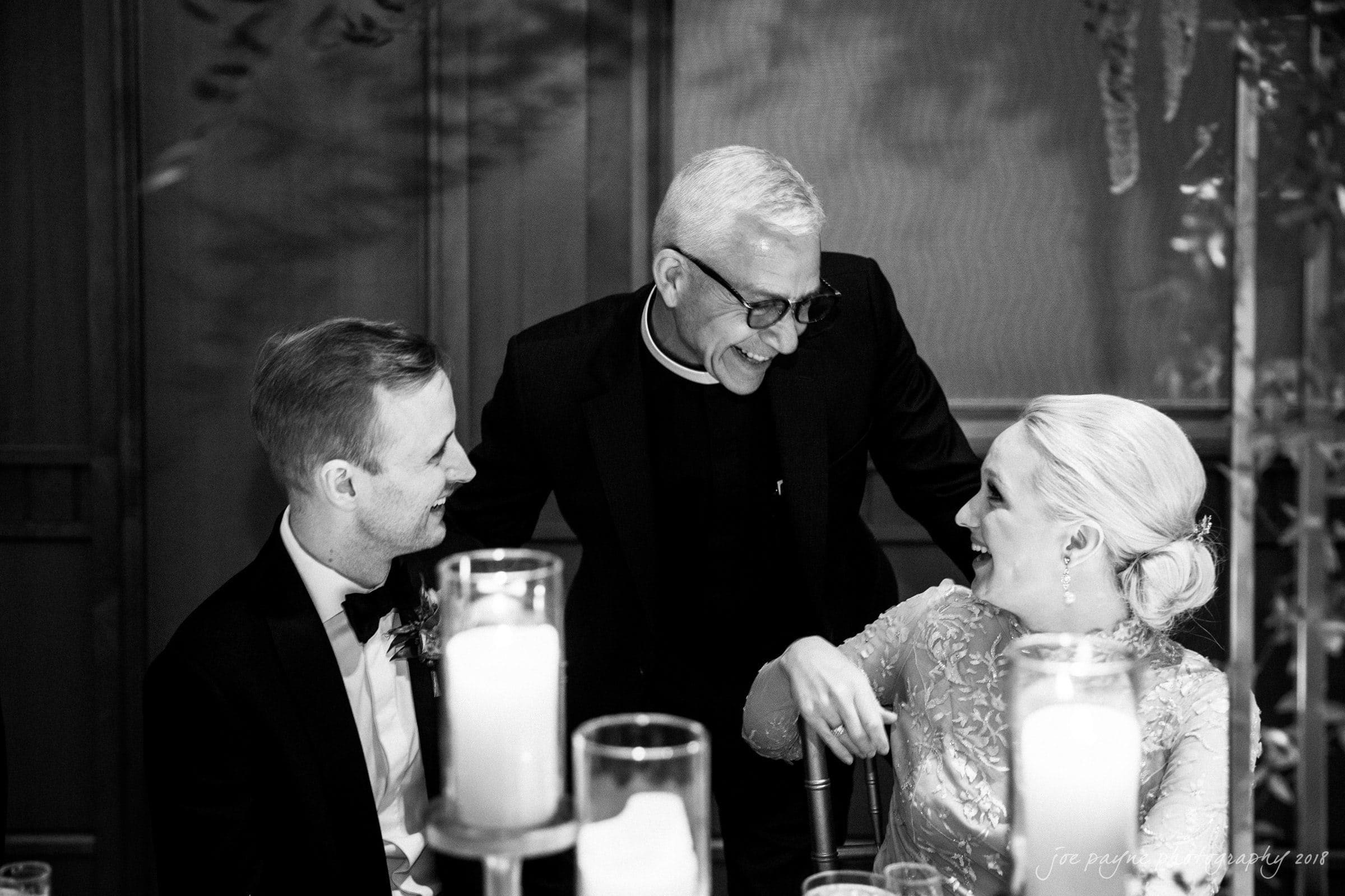 st. mary's & umstead wedding photographer - sarah & jake A black and white photograph shows a smiling couple seated at a table with lit candles, talking to a man with glasses and white hair standing beside them. The woman is wearing a fancy dress, and the man is in a suit.