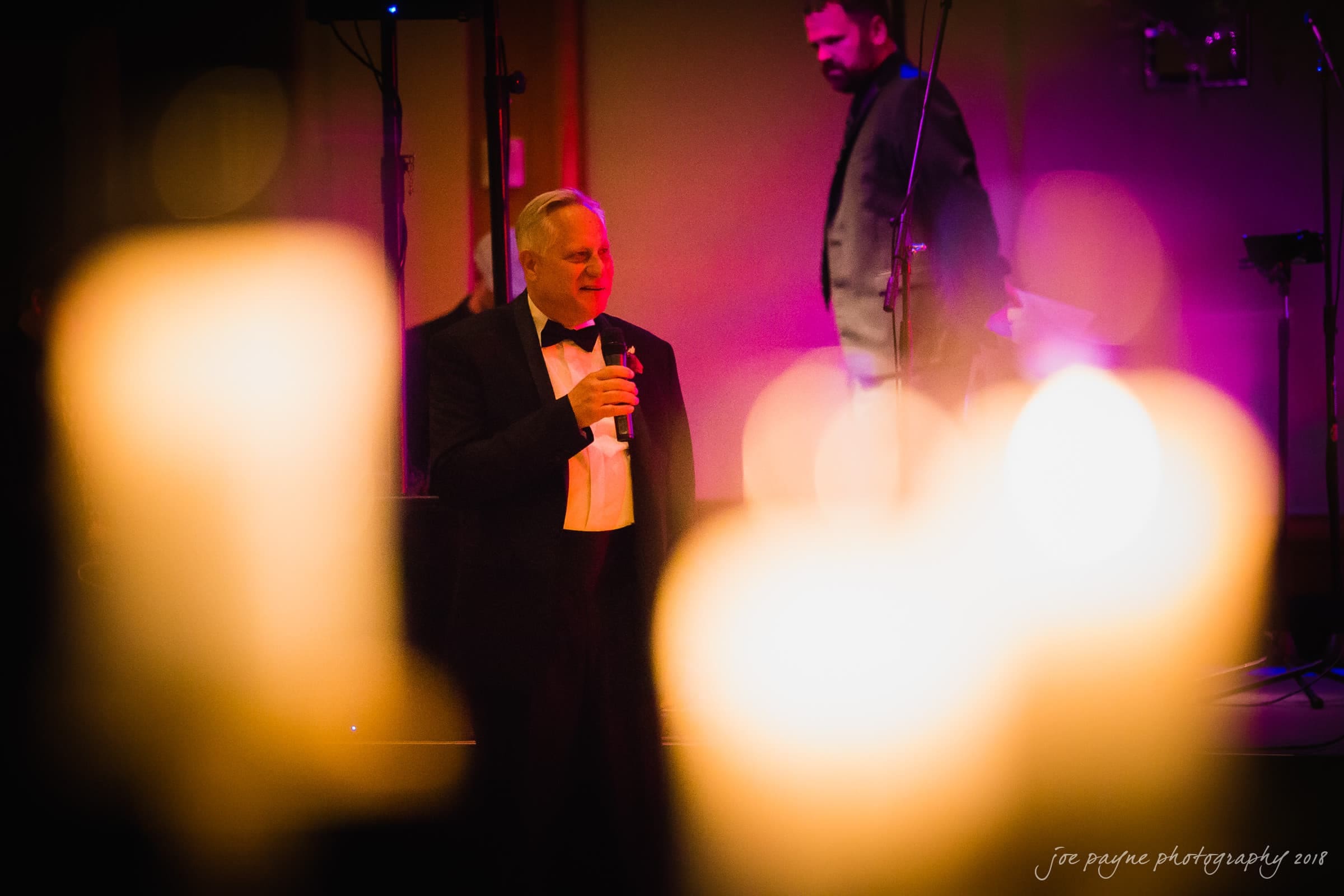 st. mary's & umstead wedding photographer - sarah & jake A man in a tuxedo holds a microphone while speaking on stage at a dimly lit event, with blurred lights in the foreground. Another man is seen in the background.