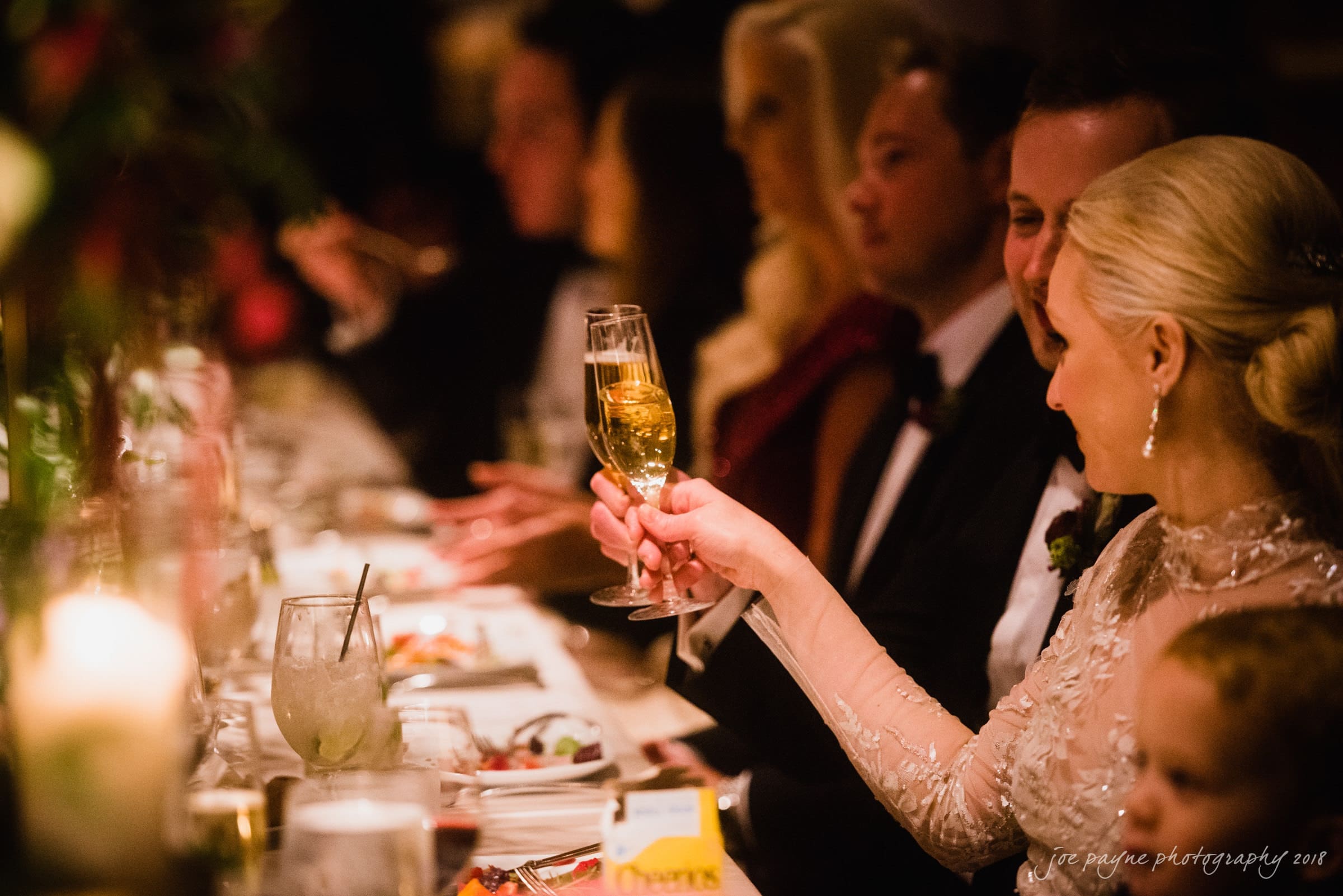 st. mary's & umstead wedding photographer - sarah & jake A bride and groom share a toast at a long dining table during a wedding reception. The focus is on their champagne glasses, and the room is warmly lit with guests seated on either side of the table.