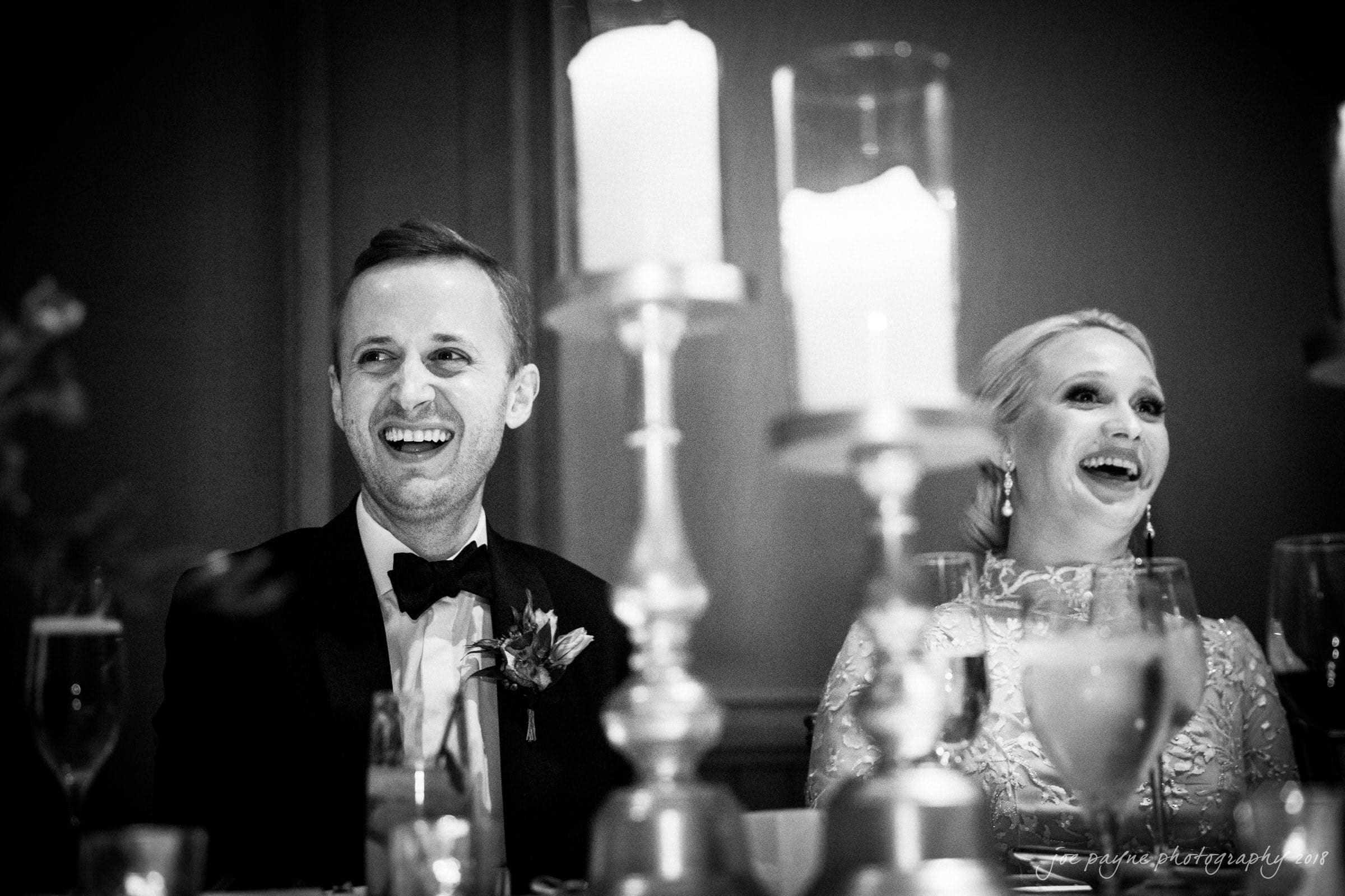 st. mary's & umstead wedding photographer - sarah & jake A black and white photo of a smiling couple seated at a table, adorned with candles and glasses. The man is in a suit with a bow tie, and the woman is in a dress with intricate detailing. Both look to their left, appearing joyful.