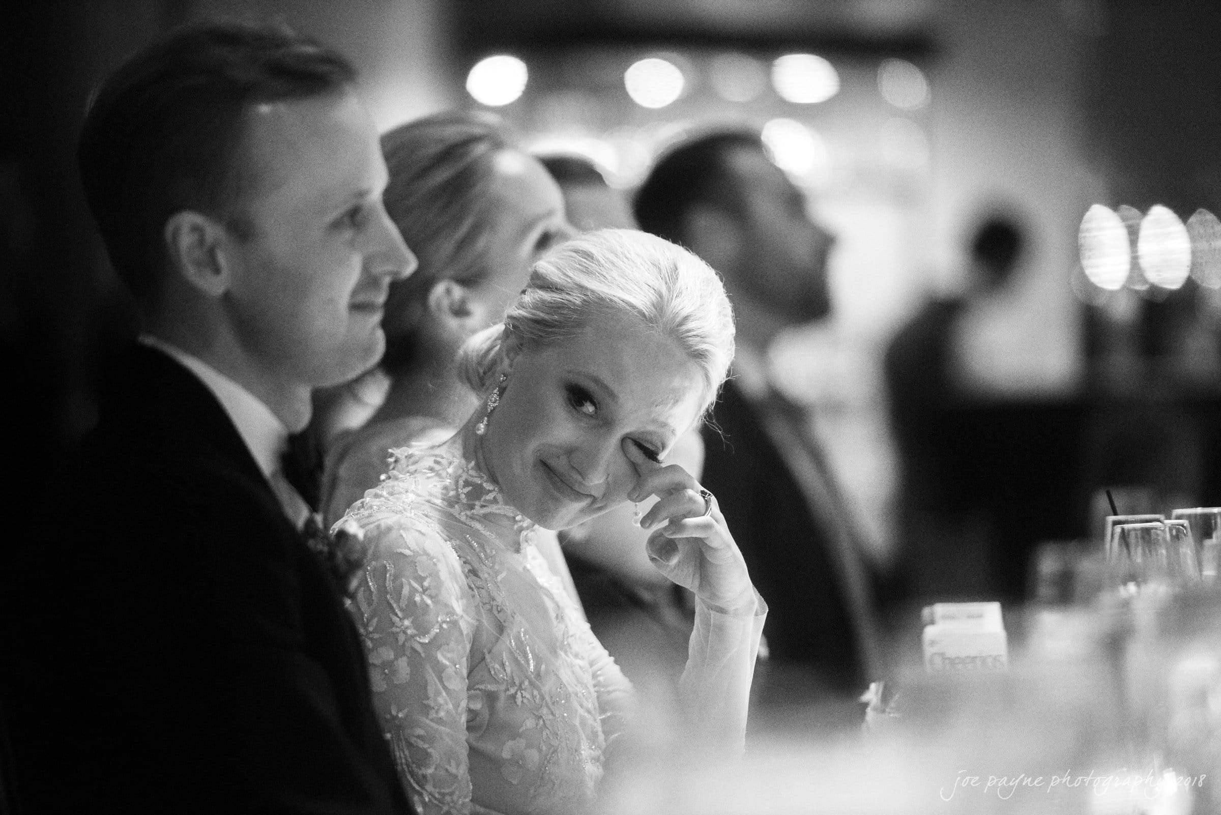 st. mary's & umstead wedding photographer - sarah & jake A bride in an elegant gown wipes a tear as she sits beside the groom at a wedding reception. Both are smiling, surrounded by guests in a softly lit, joyful setting.