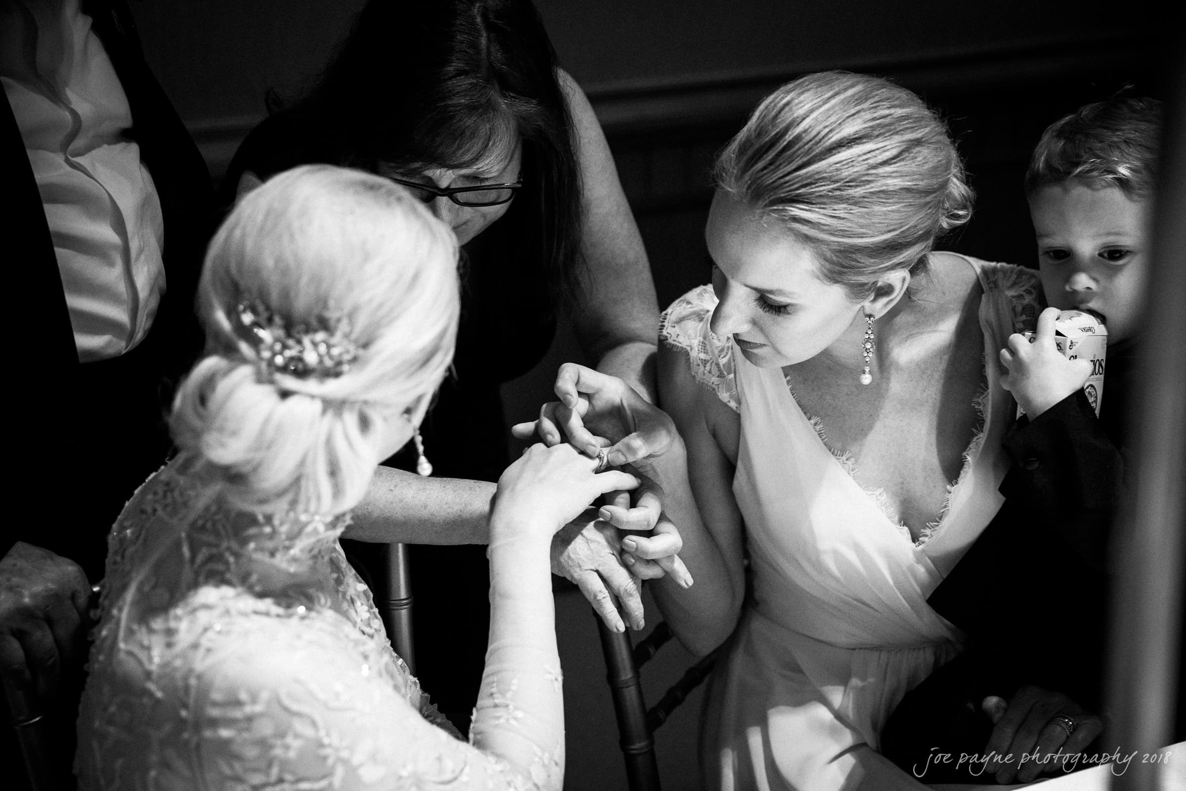 st. mary's & umstead wedding photographer - sarah & jake A bride, in an intricately beaded gown, sits as a woman in a white dress inspects her ring with a child in her arms. Another woman watches closely. The black-and-white photo captures a moment of togetherness and attention.