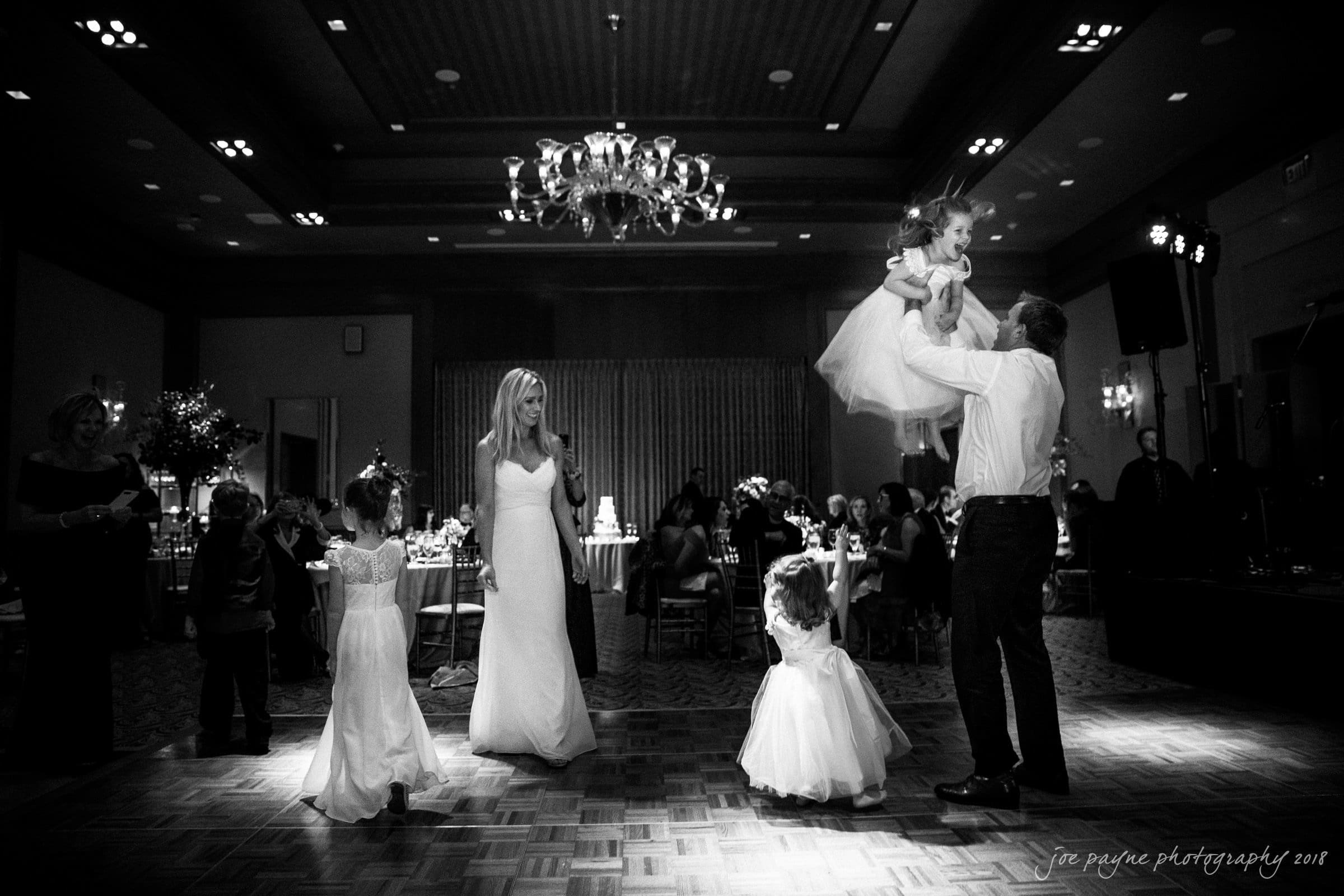 st. mary's & umstead wedding photographer - sarah & jake A black and white photo of a wedding reception with guests on the dance floor. A bride in a long dress stands smiling, while a man lifts a child in the air. Two other children, also in dresses, are nearby. The venue is elegantly decorated.