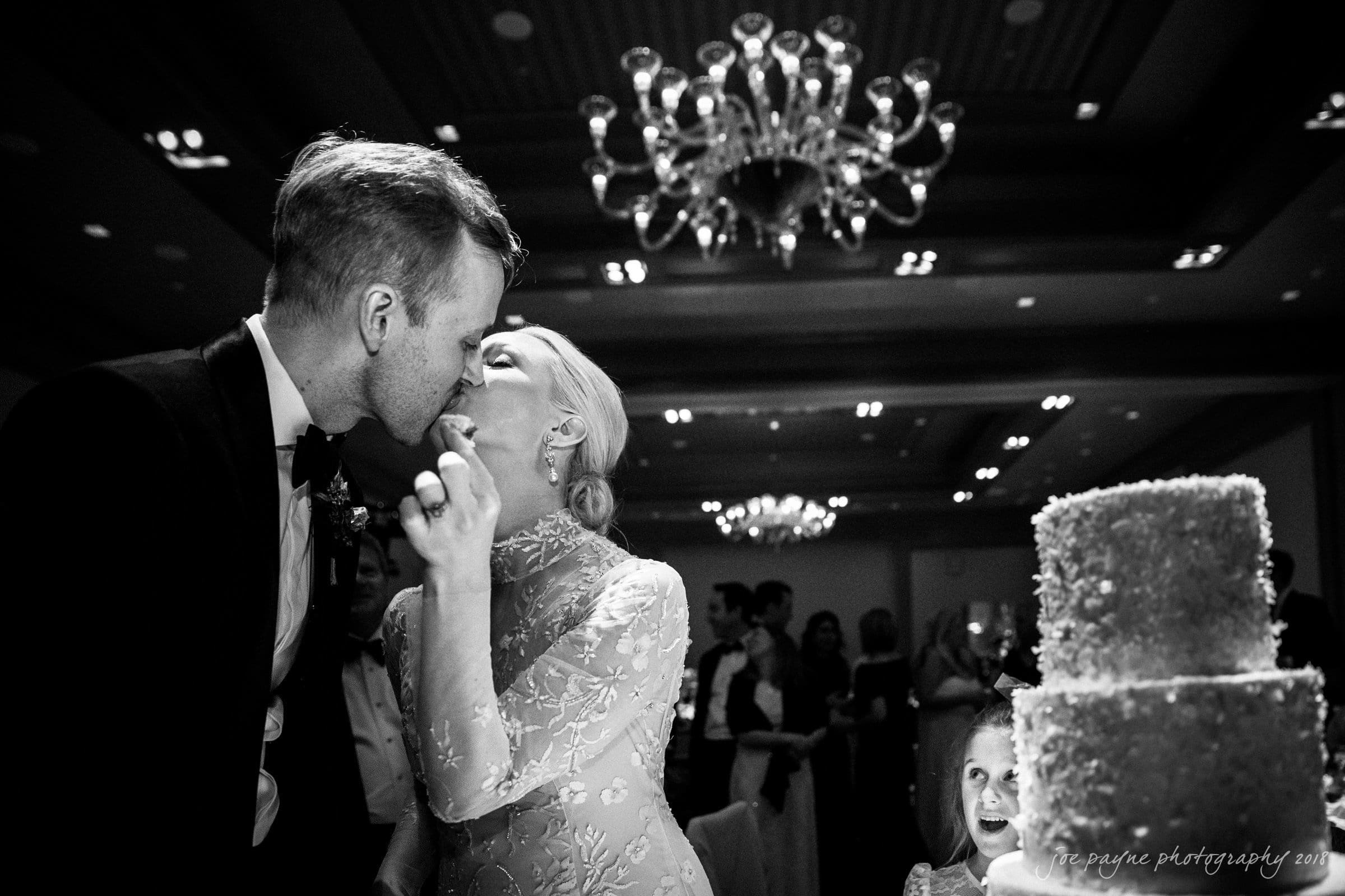st. mary's & umstead wedding photographer - sarah & jake A couple shares a kiss at their wedding reception under a chandelier. The bride wears an elegant dress, and the groom is in a suit. A tiered cake is in the foreground, and guests are visible in the background.