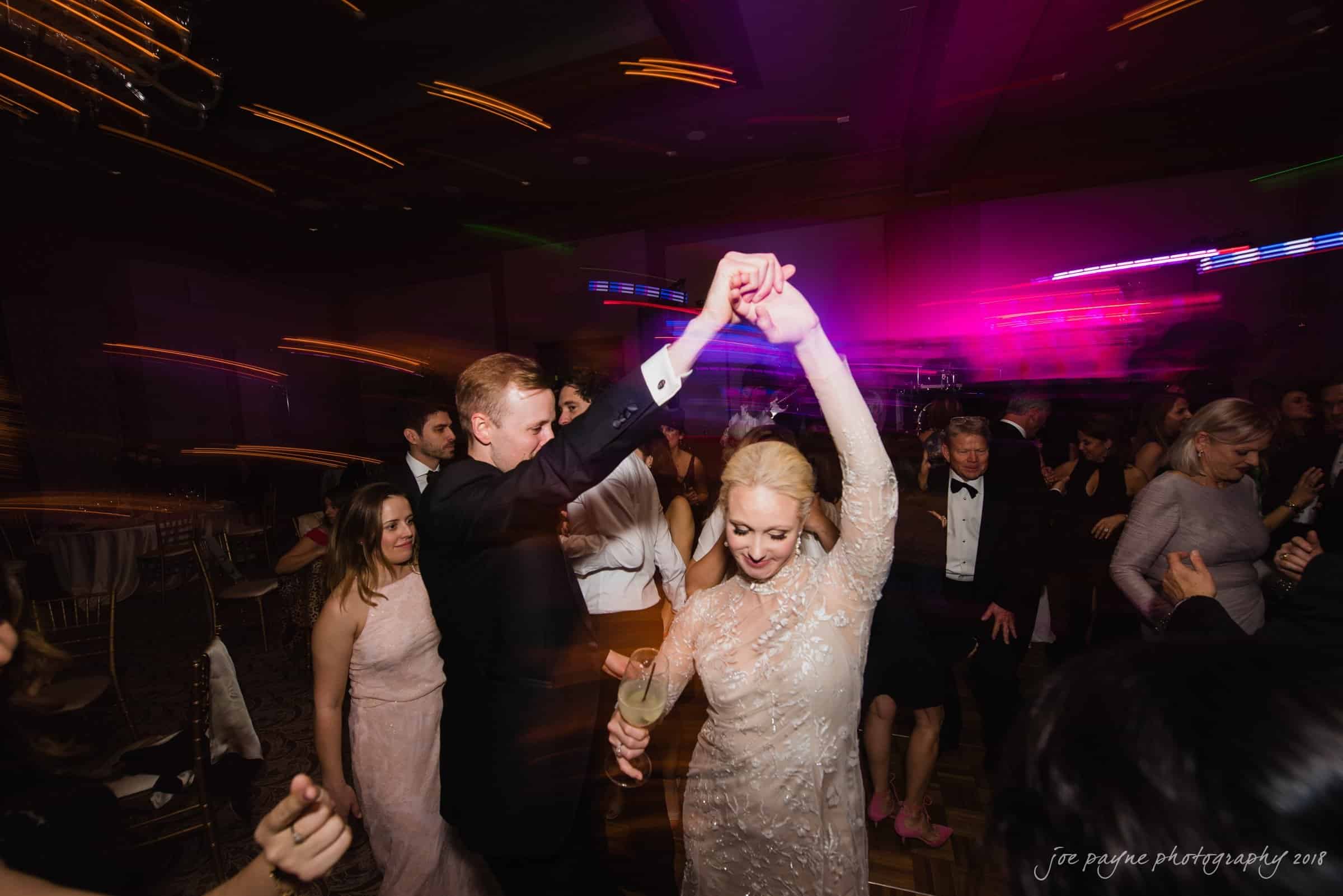 st. mary's & umstead wedding photographer - sarah & jake A couple dances joyfully at a wedding reception, surrounded by guests. The woman wears a light-colored dress, and the man is in a suit. Colorful lights blur in the background, creating a festive atmosphere.