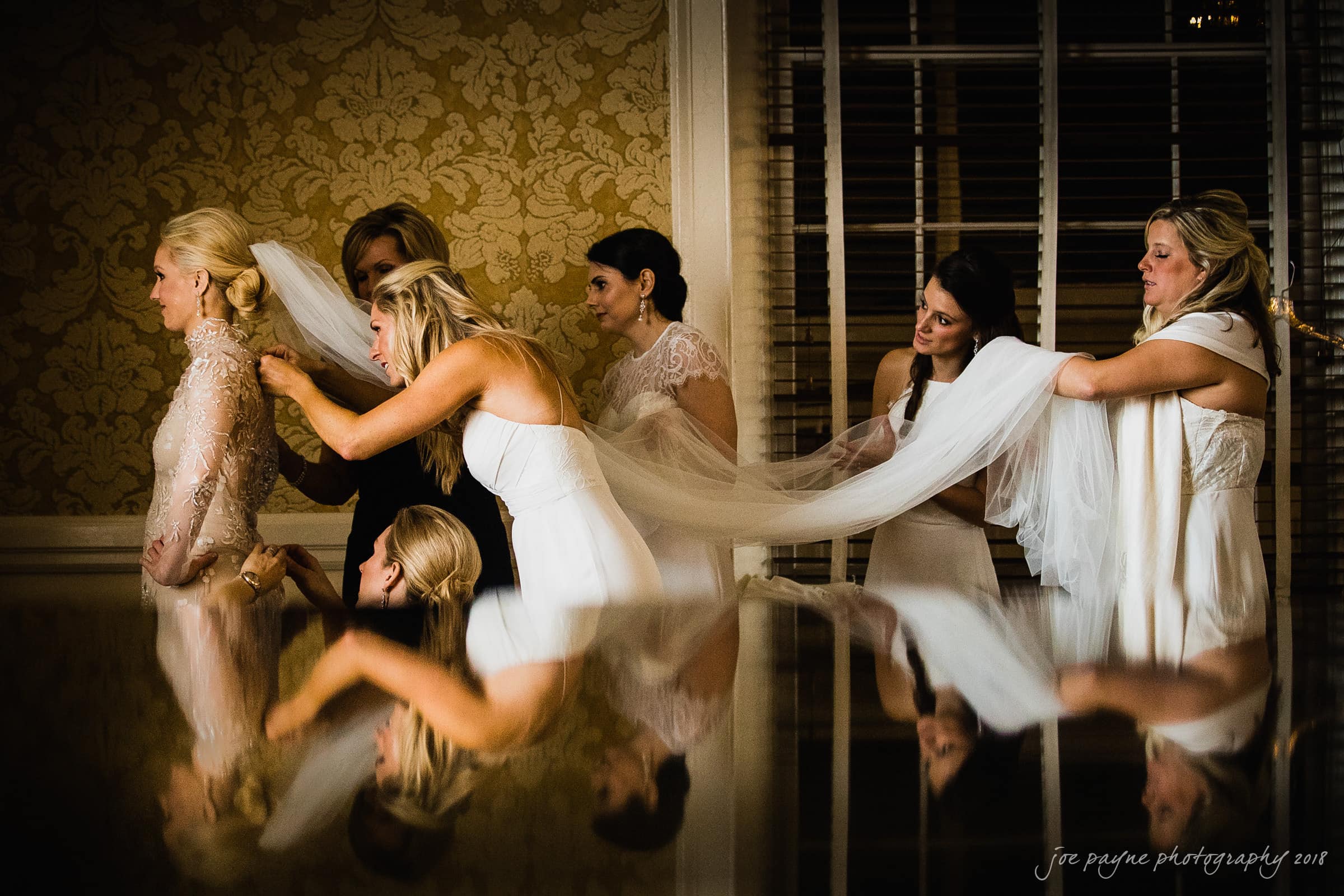 st. mary's & umstead wedding photographer - sarah & jake A bride stands in an elegant room with gold-patterned wallpaper as her bridal party assists her with the long, flowing veil. The scene is reflected on a polished surface below.