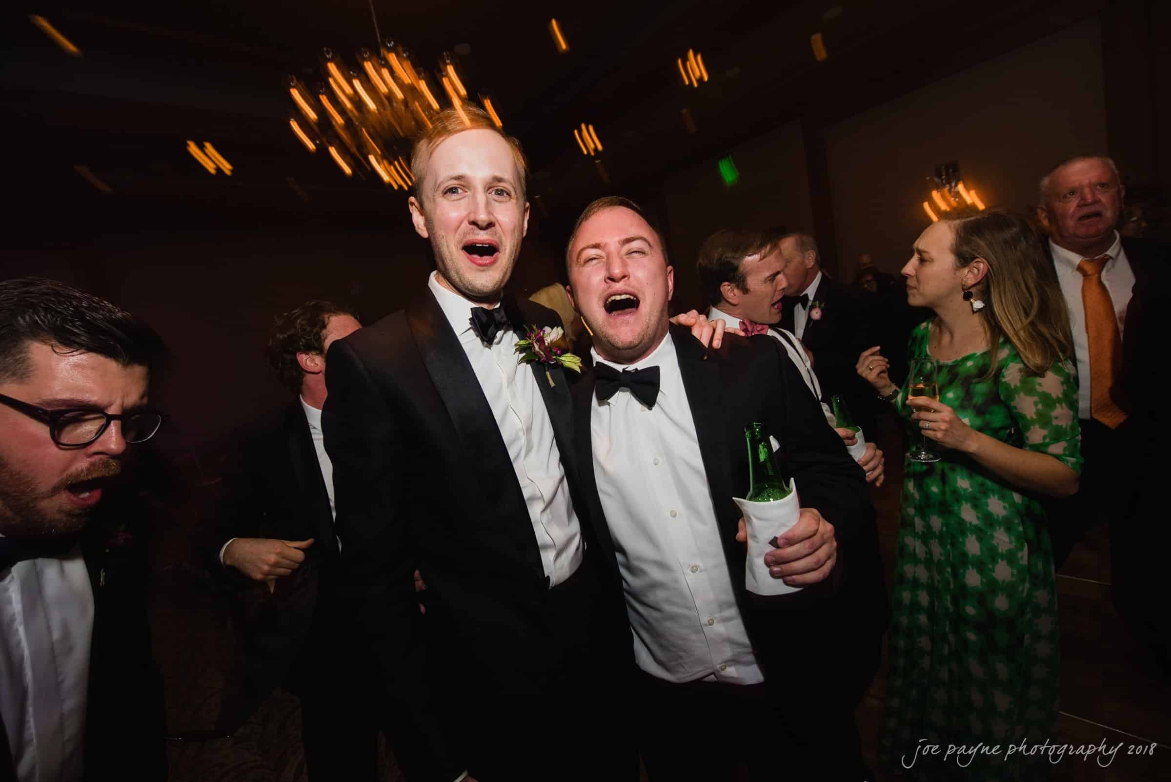 st. mary's & umstead wedding photographer - sarah & jake A group of people in formal attire are joyfully celebrating at an indoor event. Two men in the foreground, wearing tuxedos, are singing or shouting enthusiastically. A woman in a green dress and other guests are visible in the background.