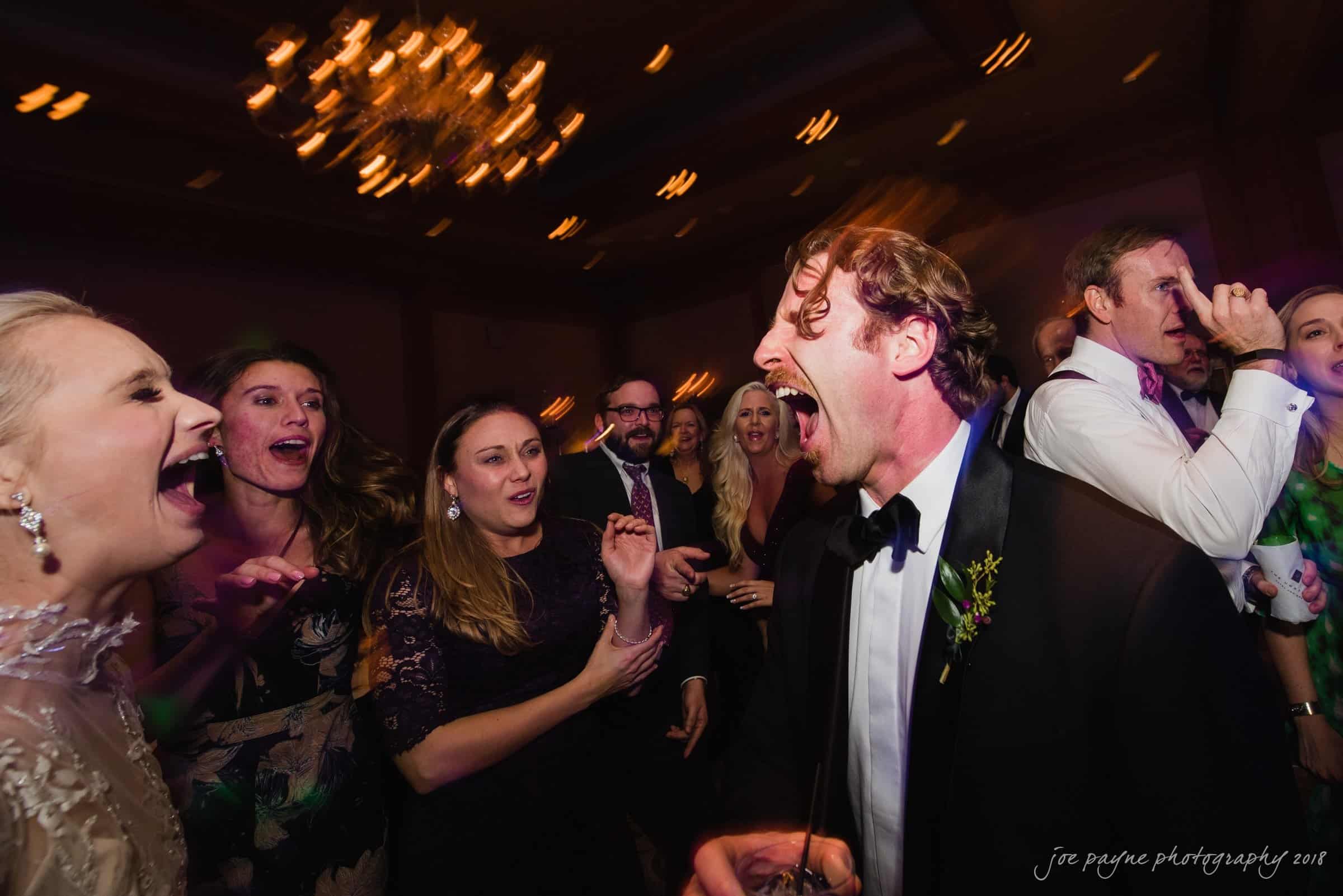 st. mary's & umstead wedding photographer - sarah & jake A joyful group of people are dancing and singing at a lively indoor event. They are dressed in formal attire, and a chandelier is visible in the background, casting a warm glow over the scene.