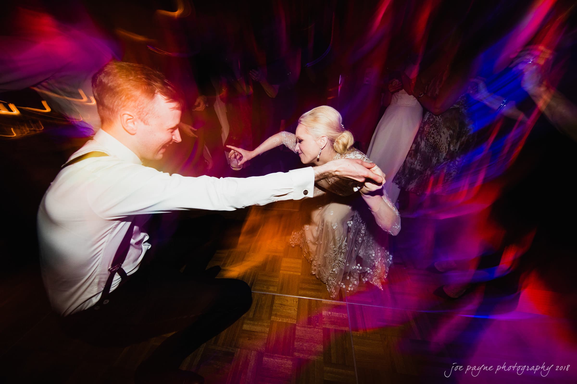 st. mary's & umstead wedding photographer - sarah & jake A man and woman dance energetically on a dimly lit dance floor, surrounded by vibrant streaks of colorful lights. The woman is in a flowing dress, and the man is in formal attire. The scene captures movement and celebration.