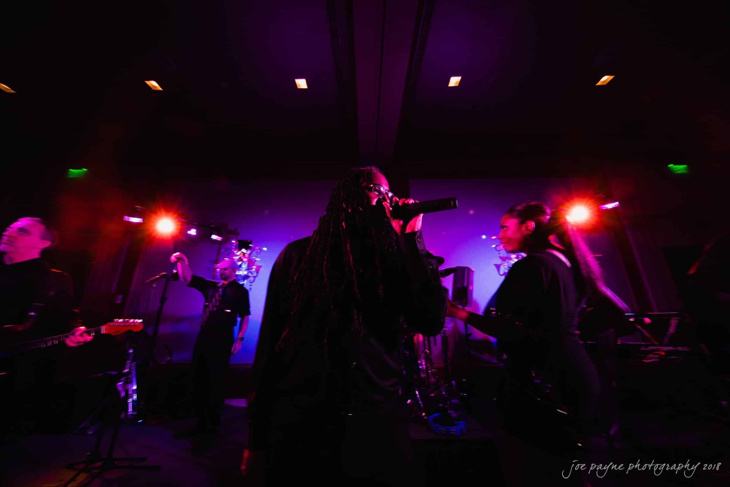 st. mary's & umstead wedding photographer - sarah & jake A band performs energetically on stage under purple and pink lights. The focus is on a singer holding a microphone, surrounded by musicians. The atmosphere is vibrant and dynamic, capturing the essence of a live music event.