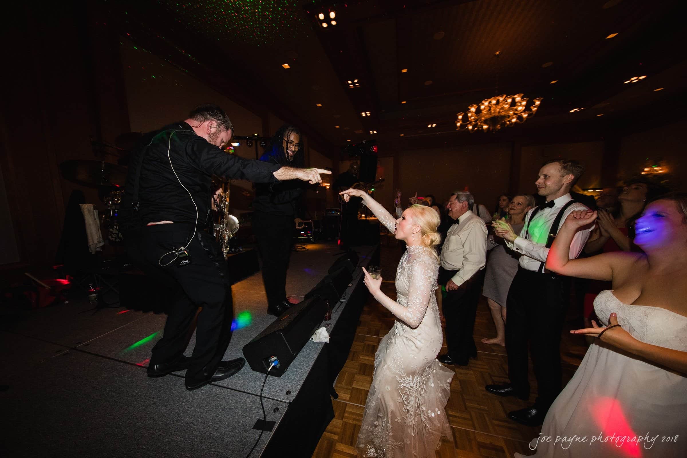 st. mary's & umstead wedding photographer - sarah & jake A lively wedding reception with a bride and guests dancing enthusiastically near a stage. One guest interacts with a musician, while others cheerfully join in the celebration. The room is warmly lit with elegant chandeliers.