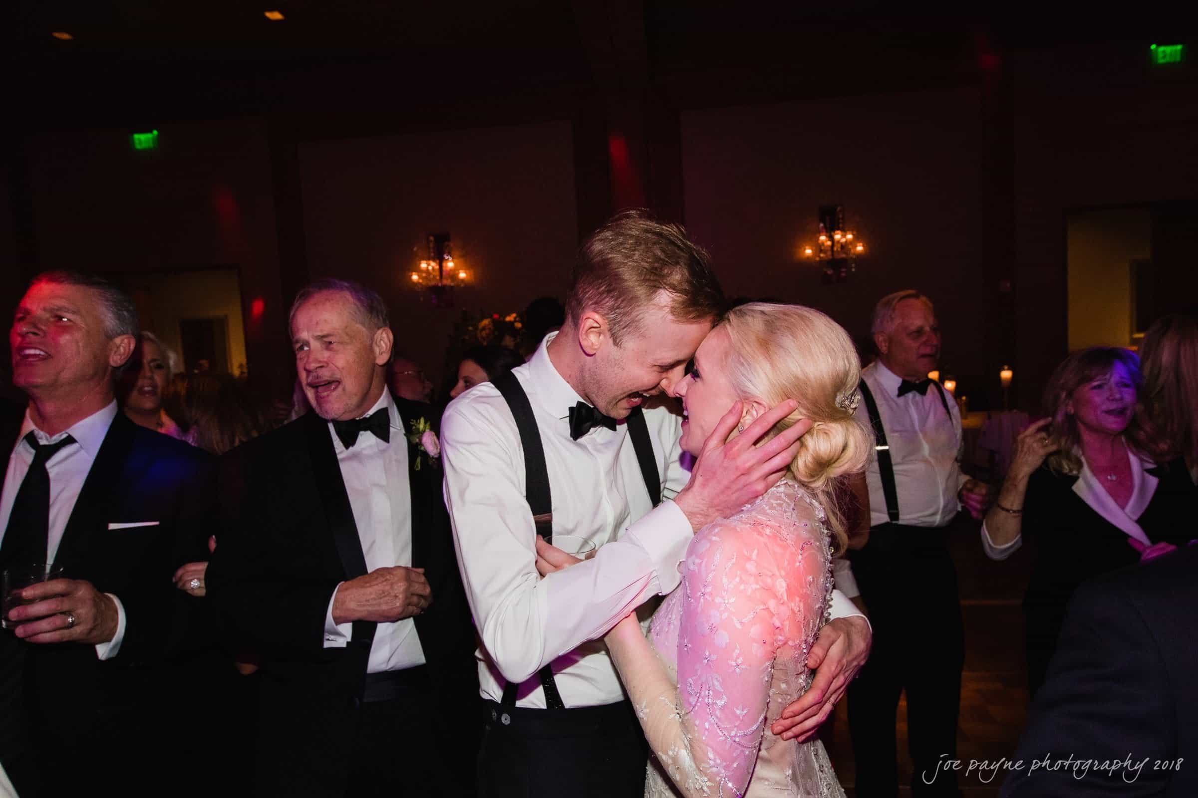 st. mary's & umstead wedding photographer - sarah & jake A couple embraces joyfully on a dance floor, surrounded by guests in formal attire. The setting is dimly lit, creating a warm atmosphere. Laughter and excitement fill the air as others dance and celebrate around them.