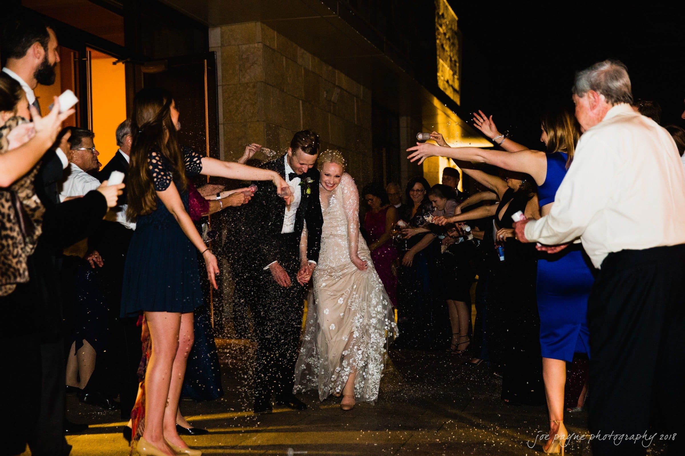 st. mary's & umstead wedding photographer - sarah & jake A newlywed couple walks through a crowd of guests at night. The bride and groom are showered with confetti as they smile and hold hands. Guests at both sides cheer and celebrate under warm lighting.