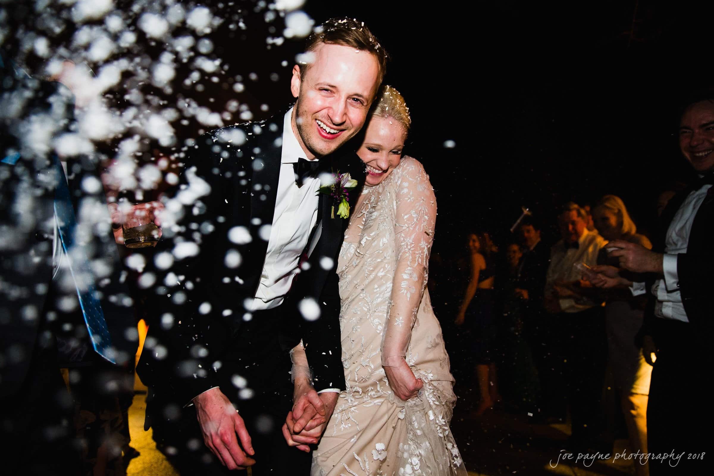 st. mary's & umstead wedding photographer - sarah & jake A smiling couple in formal attire is showered with confetti as they walk arm in arm. The woman wears a lace dress, and the man is in a tuxedo. People are gathered in the background, celebrating at night.