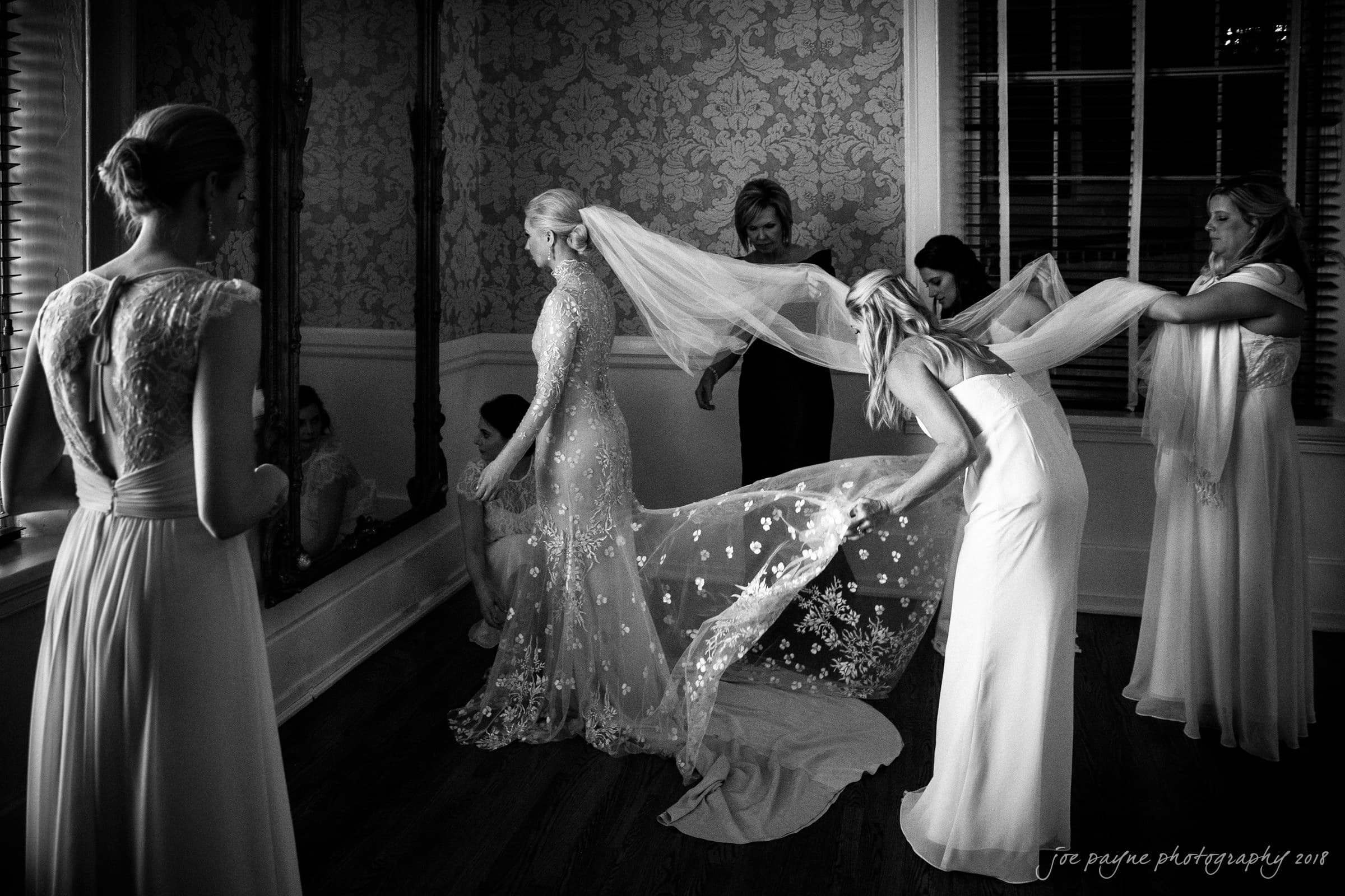 st. mary's & umstead wedding photographer - sarah & jake A bride in a flowing gown stands near a window as bridesmaids assist with her veil in a dimly lit room. Large mirrors and patterned wallpaper create an elegant, intimate atmosphere.