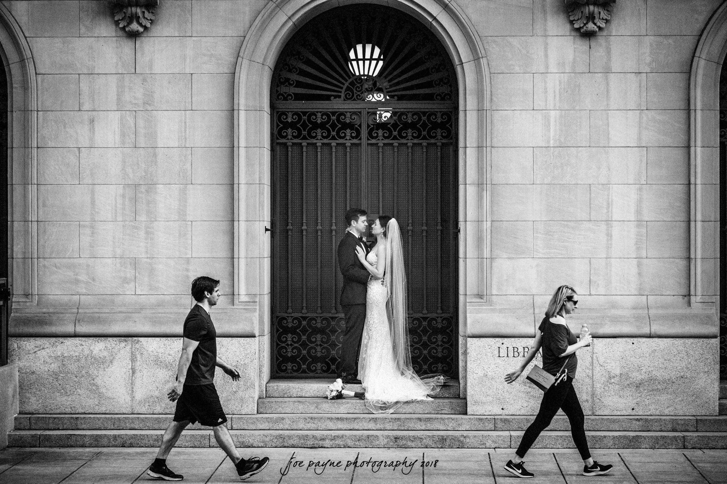 A black-and-white photo of a bride and groom kissing in front of a gated entrance. The bride wears a long veil and gown. Two pedestrians, one man and one woman, walk past them in opposite directions. The setting appears urban, with a stone building.
