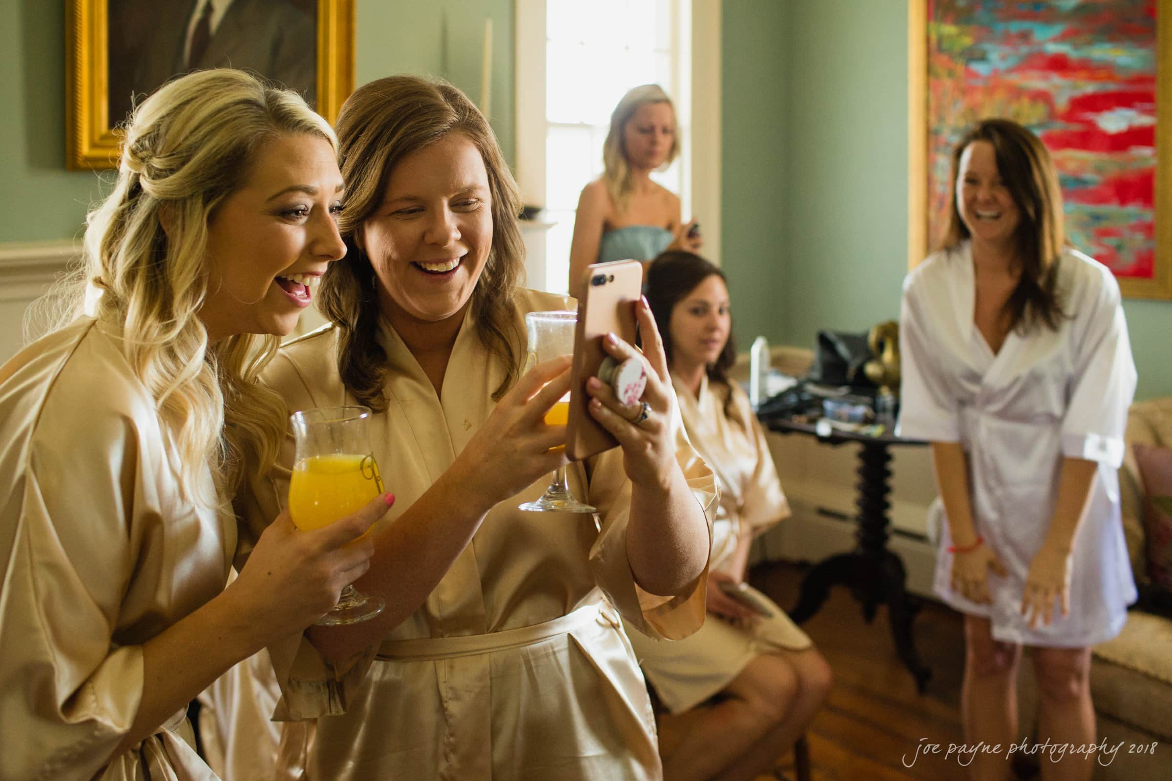 may-lew farm wedding photography - macy & jordan A group of women wearing light-colored robes are in a room. Two women in the foreground are smiling and holding drinks, looking at a phone. Others in the background appear to be talking and laughing. The atmosphere is joyful and relaxed.