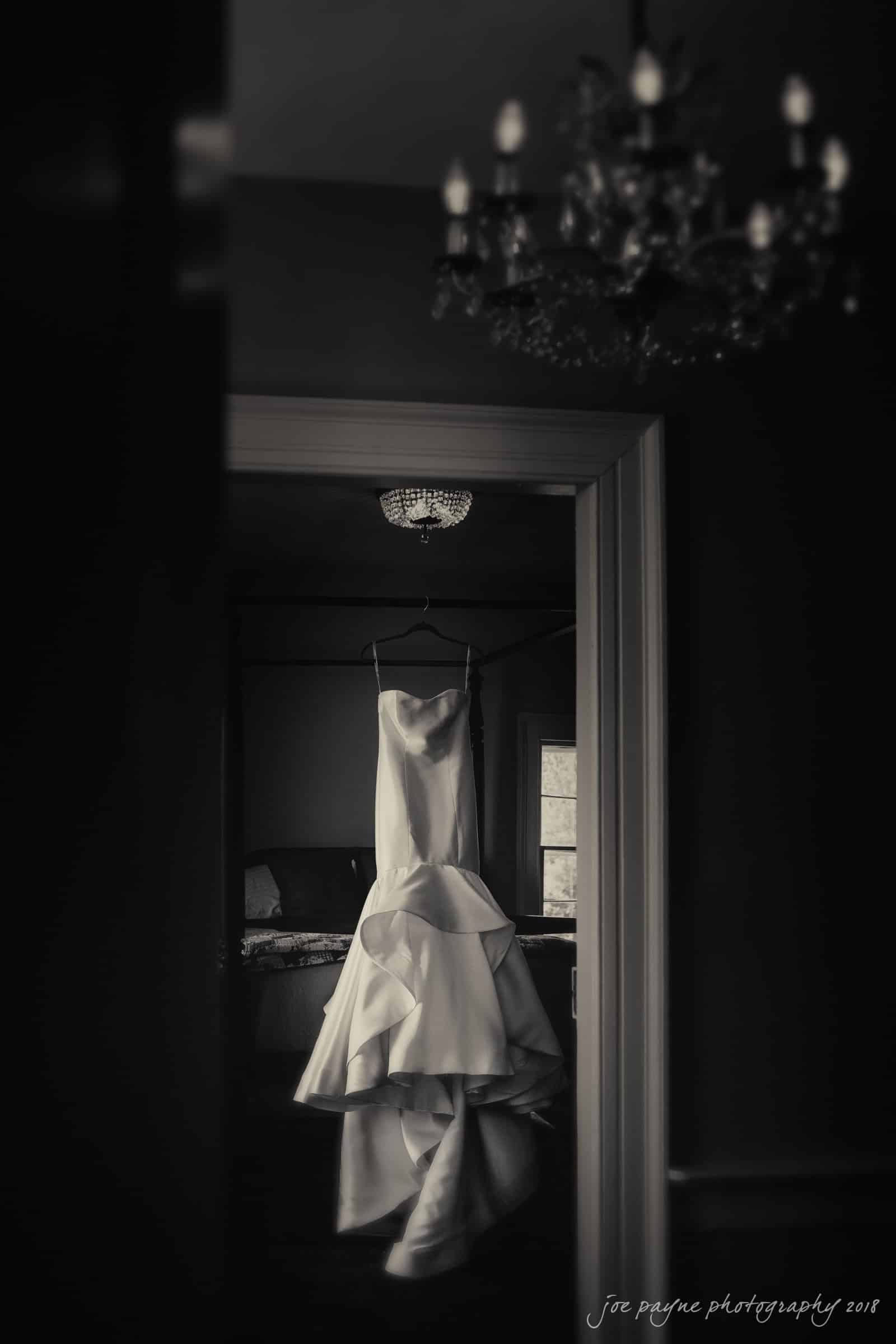 may-lew farm wedding photography - macy & jordan A black and white photo of a strapless, full-length wedding gown hanging in a dimly lit room. The dress is elegantly displayed on a hanger with soft, flowing fabric. A chandelier is visible on the ceiling, adding an elegant touch.