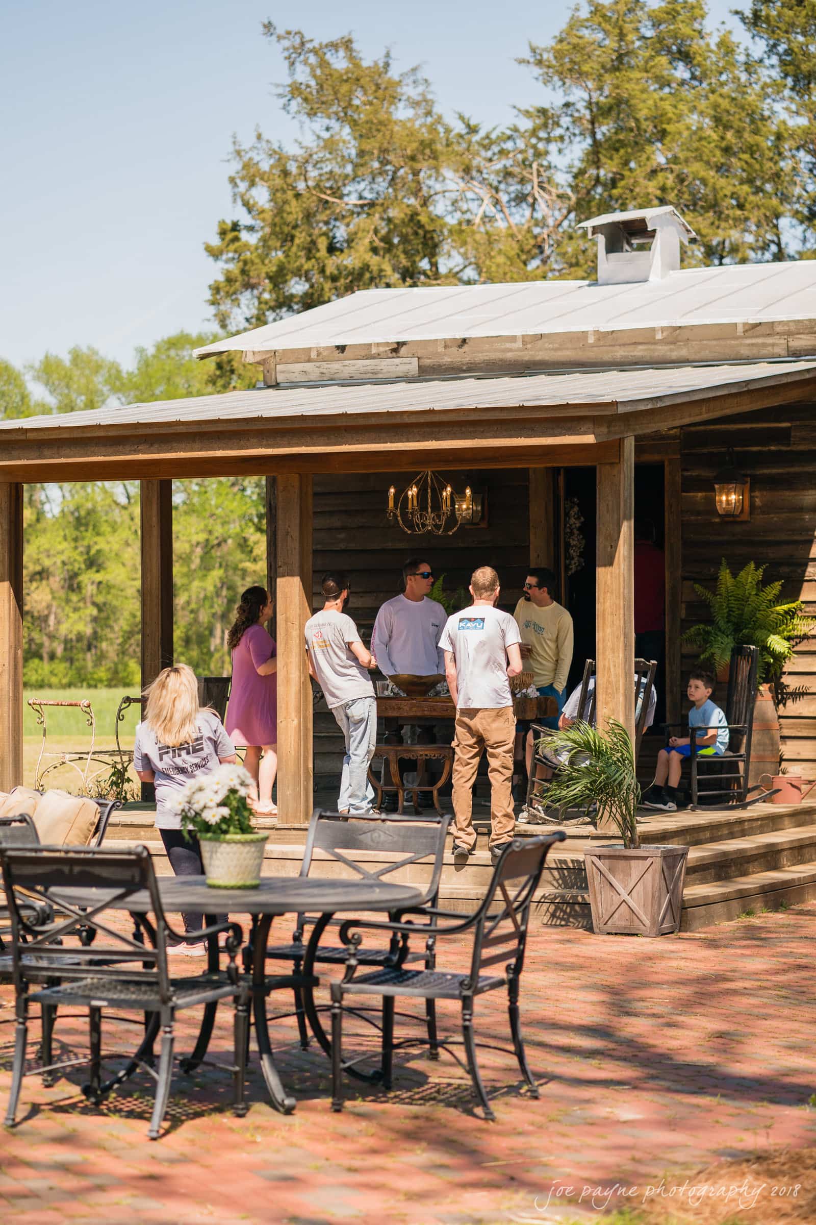 may-lew farm wedding photography - macy & jordan A group of people socializing under a wooden pavilion in a sunny outdoor setting. In the foreground, there are metal chairs and a table with a potted plant on a brick patio. Trees and greenery are visible in the background.