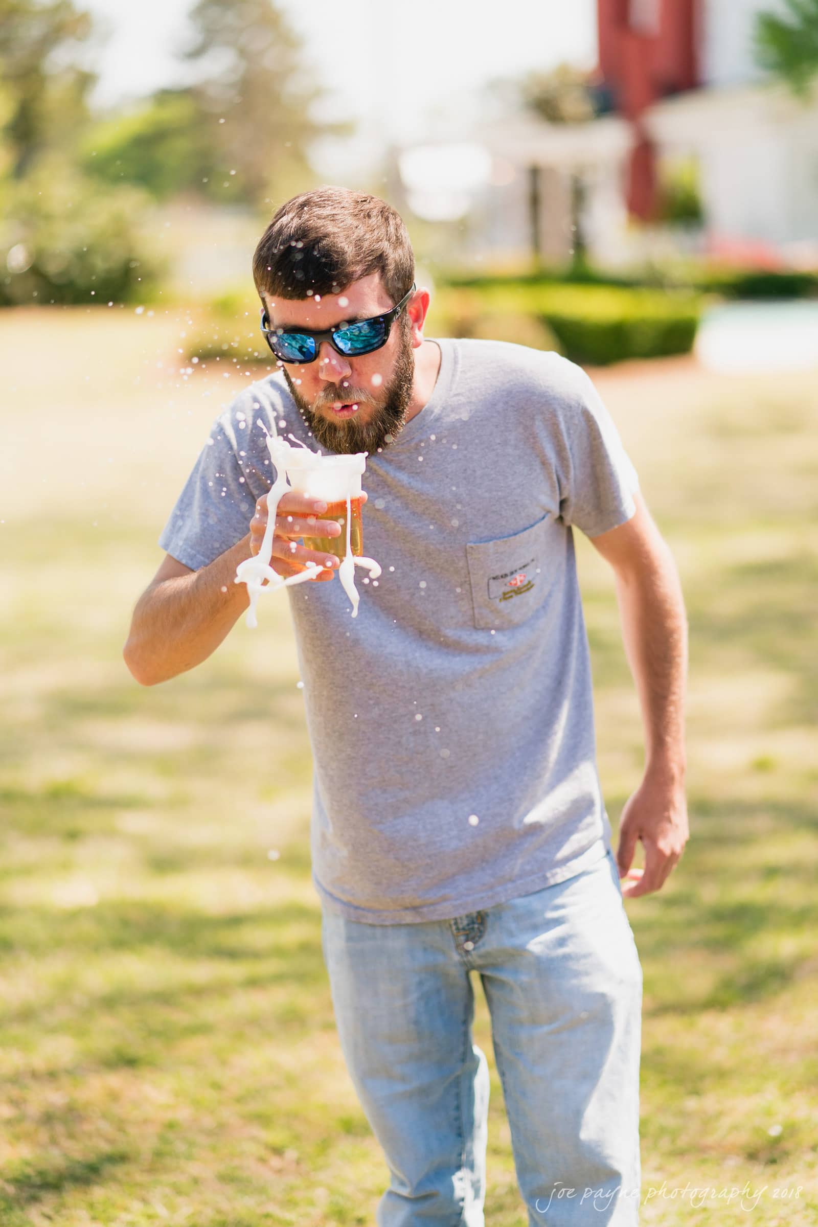 may-lew farm wedding photography - macy & jordan A bearded man wearing sunglasses accidentally spills his drink while walking on a lawn. The liquid splashes mid-air as he tries to catch it. He is casually dressed in a gray t-shirt and jeans. A house and greenery are in the background.