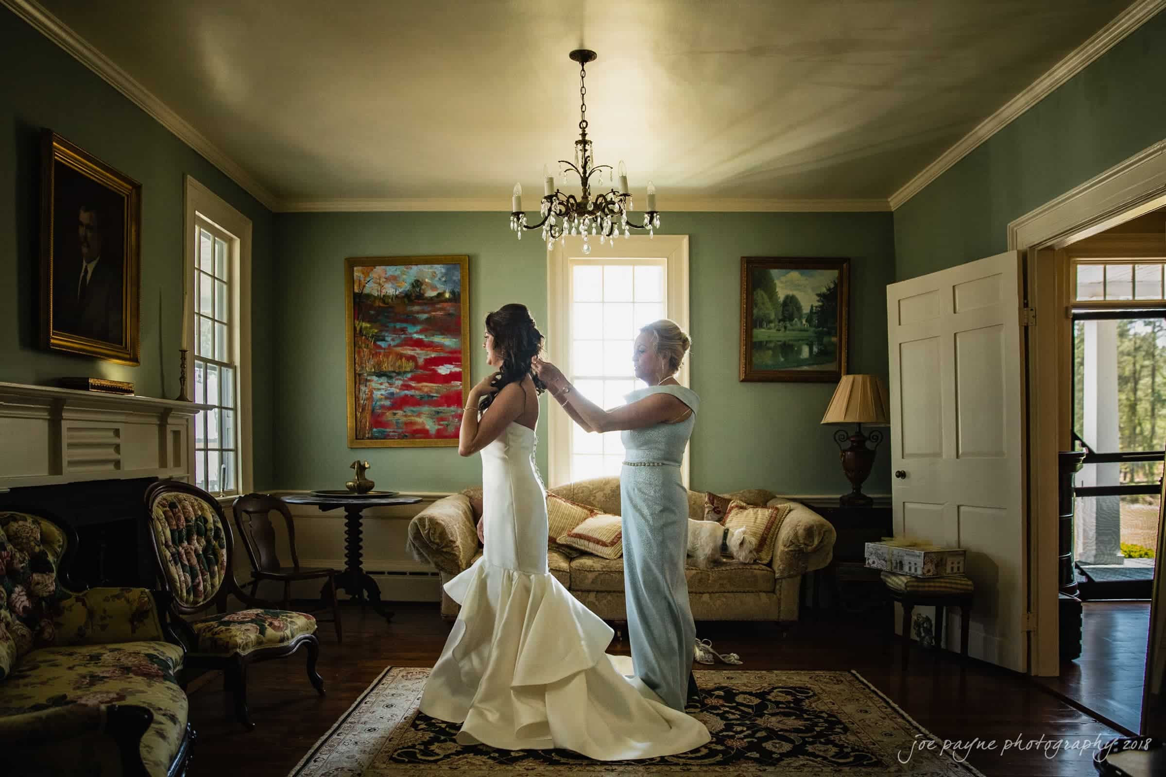may-lew farm wedding photography - macy & jordan A bride in a white gown stands in a vintage-decorated room, while another woman adjusts her hair. Sunlight streams in through the windows, and paintings adorn the walls. A chandelier hangs above them.