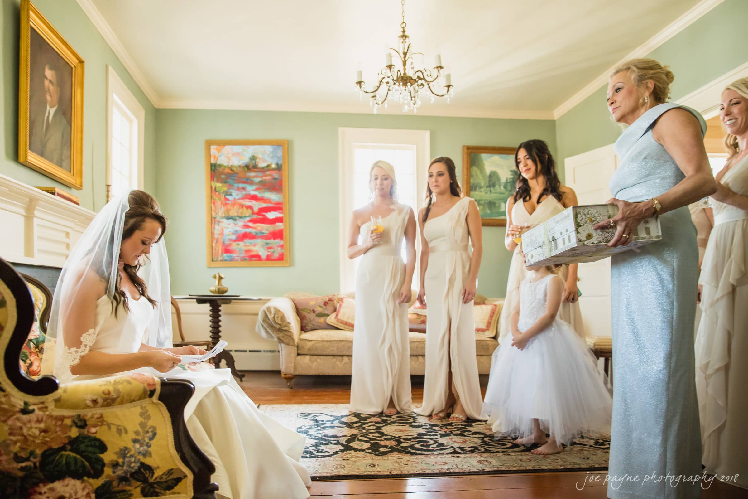 may-lew farm wedding photography - macy & jordan A bride in a white dress sits reading a letter, surrounded by bridesmaids in matching gowns. An older woman holds a decorative box. The room is elegant, with paintings and a chandelier.